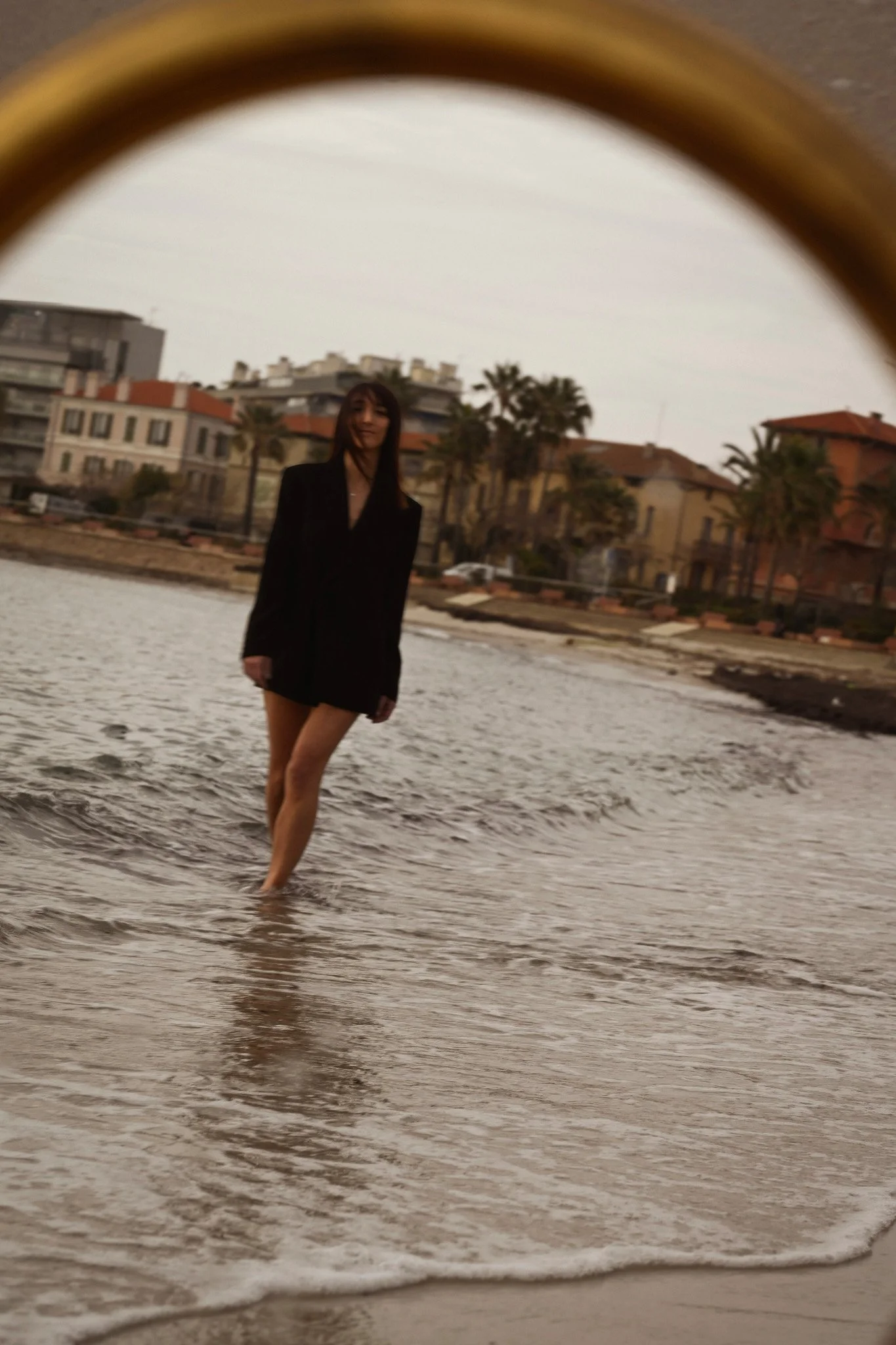 Une femme en veste noire, debout dans l'eau, avec des bâtiments résidentiels et des palmiers en arrière-plan, vue à travers une surface courbée qui ressemble à une lentille ou un miroir.