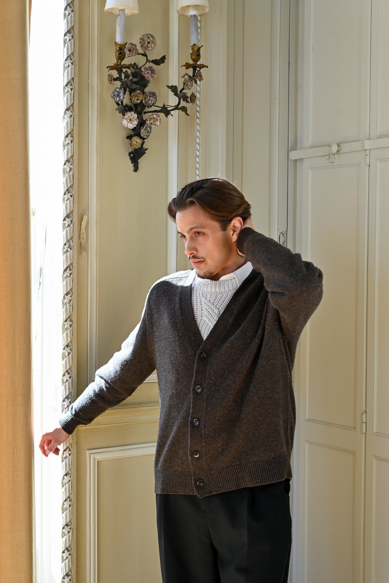 Jeune homme en pull gris et cardigan marron, se tenant près d'une fenêtre, se touchant la nuque, dans une pièce lumineuse avec des murs clair et un décor floral.