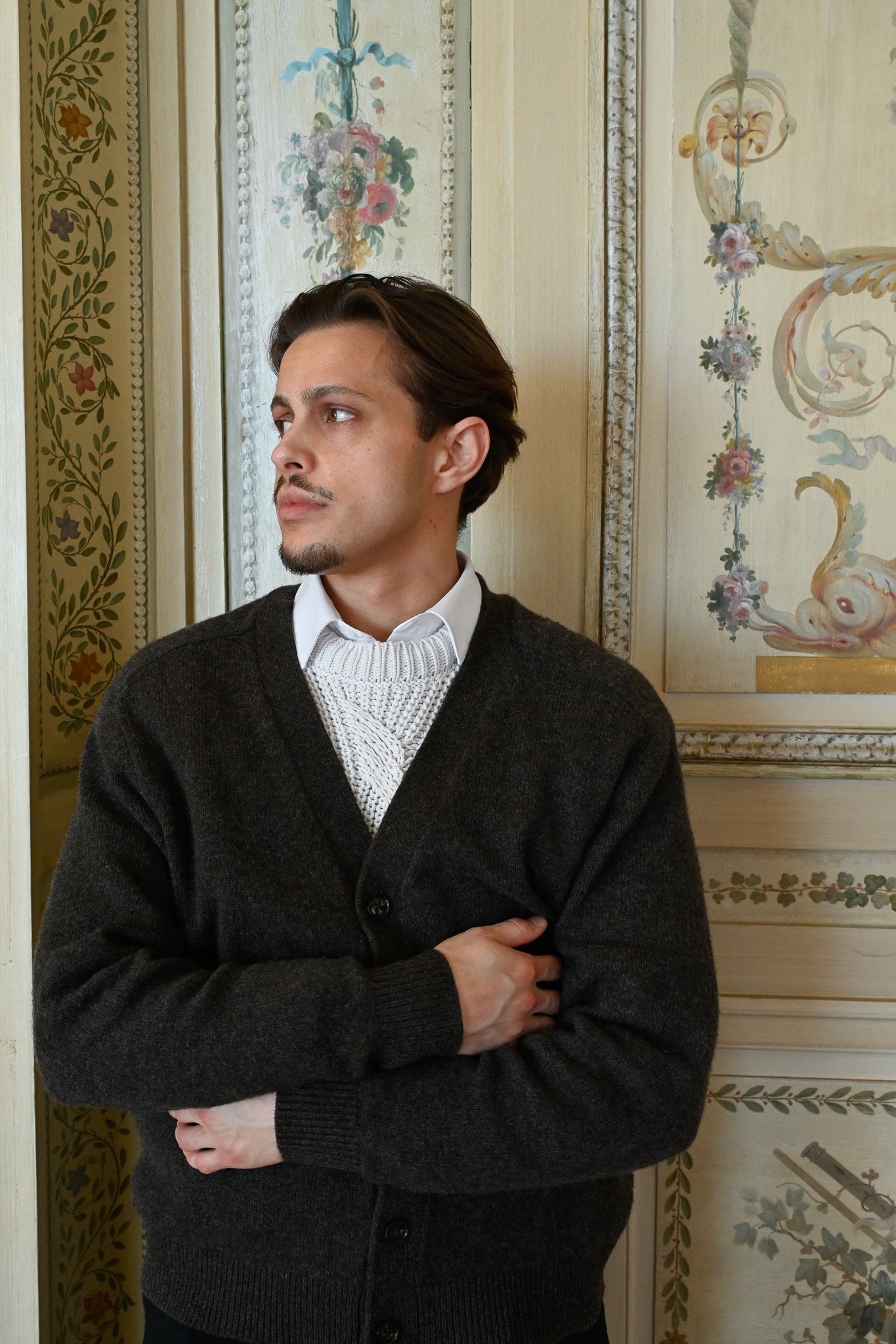 Un homme avec des cheveux bruns, portant un pull gris foncé et une chemise blanche, se tient devant un mur décoratif avec des motifs floraux et des ornements élégants.