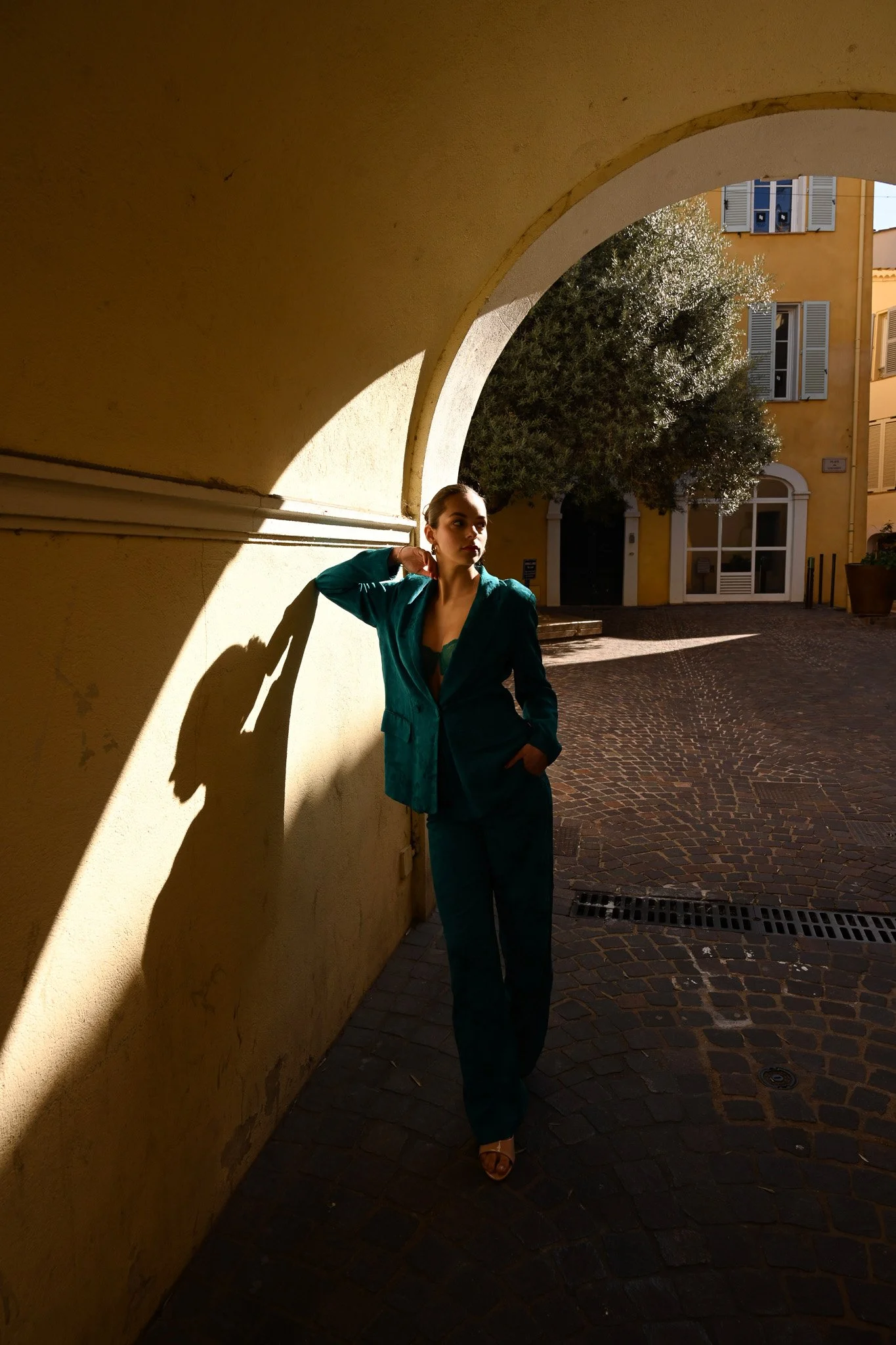 Une femme en tailleur bleu se tient devant un mur jaune sous un arc, en plein soleil, avec un arbre vert en arrière-plan.
