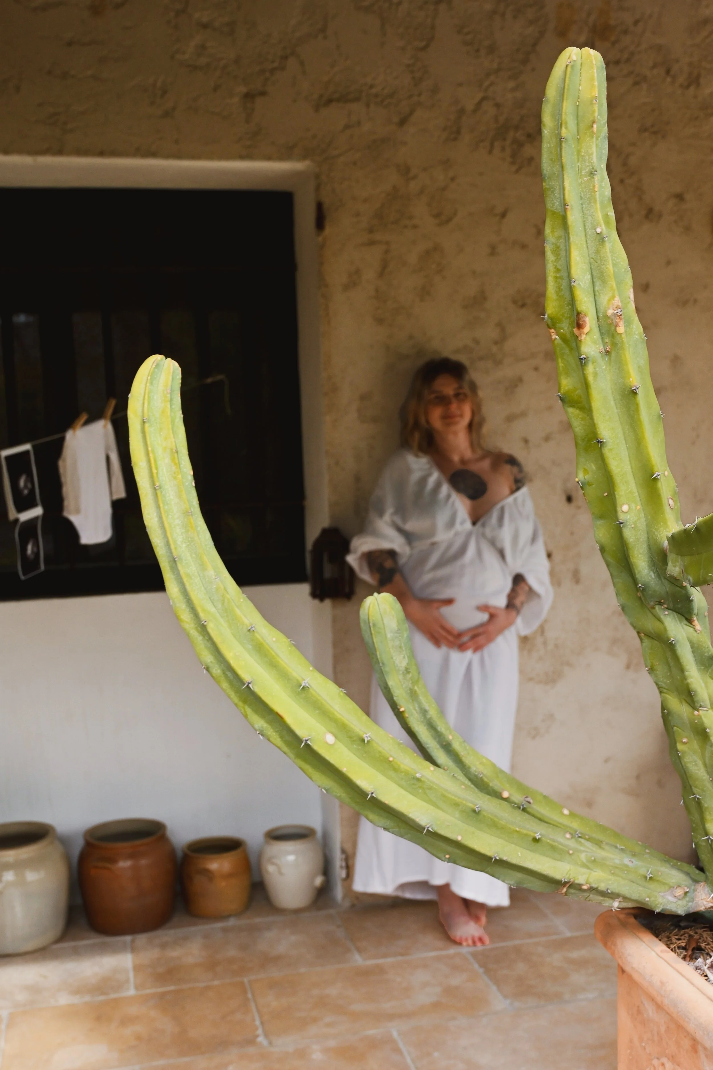 Une femme enceinte portant une robe blanche se tient derrière un cactus vert placé en avant-plan dans une pièce avec des pots de décoration en arrière-plan.