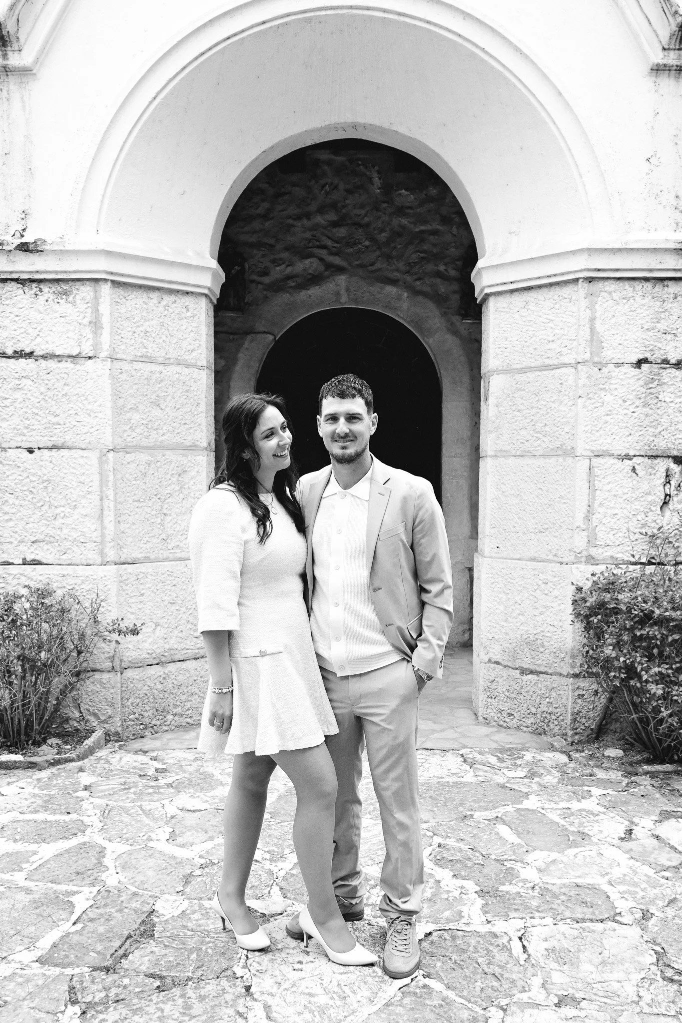 Un couple habillé en blanc se tenant devant une arche en pierre, se souriant, en extérieur.
