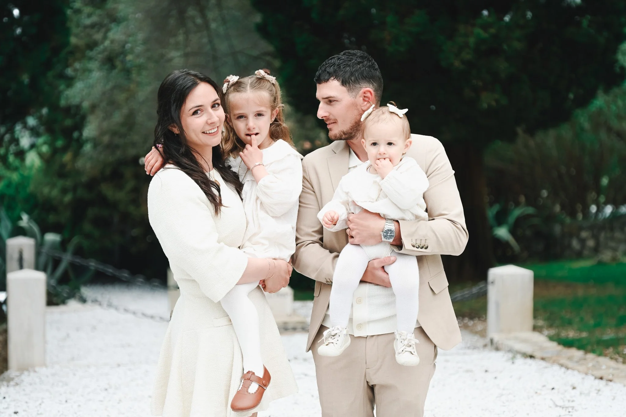 Une famille heureuse composée de deux adultes et deux jeunes enfants, en tenue claire, se promène dans un parc verdoyant. La mère porte une robe blanche, le père un costume beige, tandis que les enfants portent également des vêtements blancs. La scèn