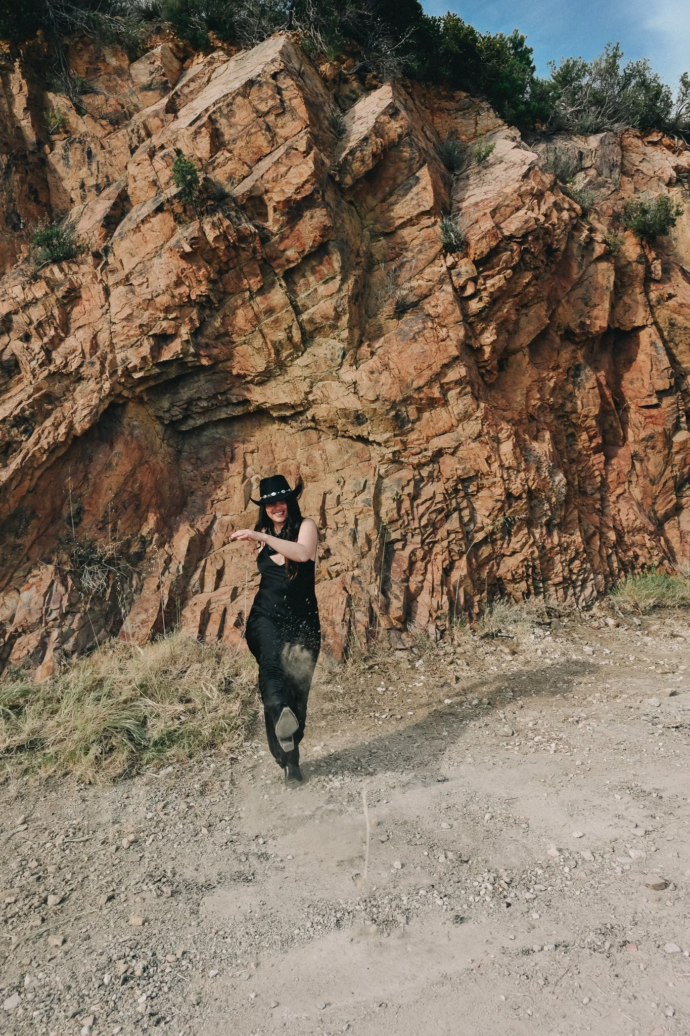 Une femme habillée en noir avec un chapeau noir danse ou saute devant un mur de roches rouges et un ciel bleu.