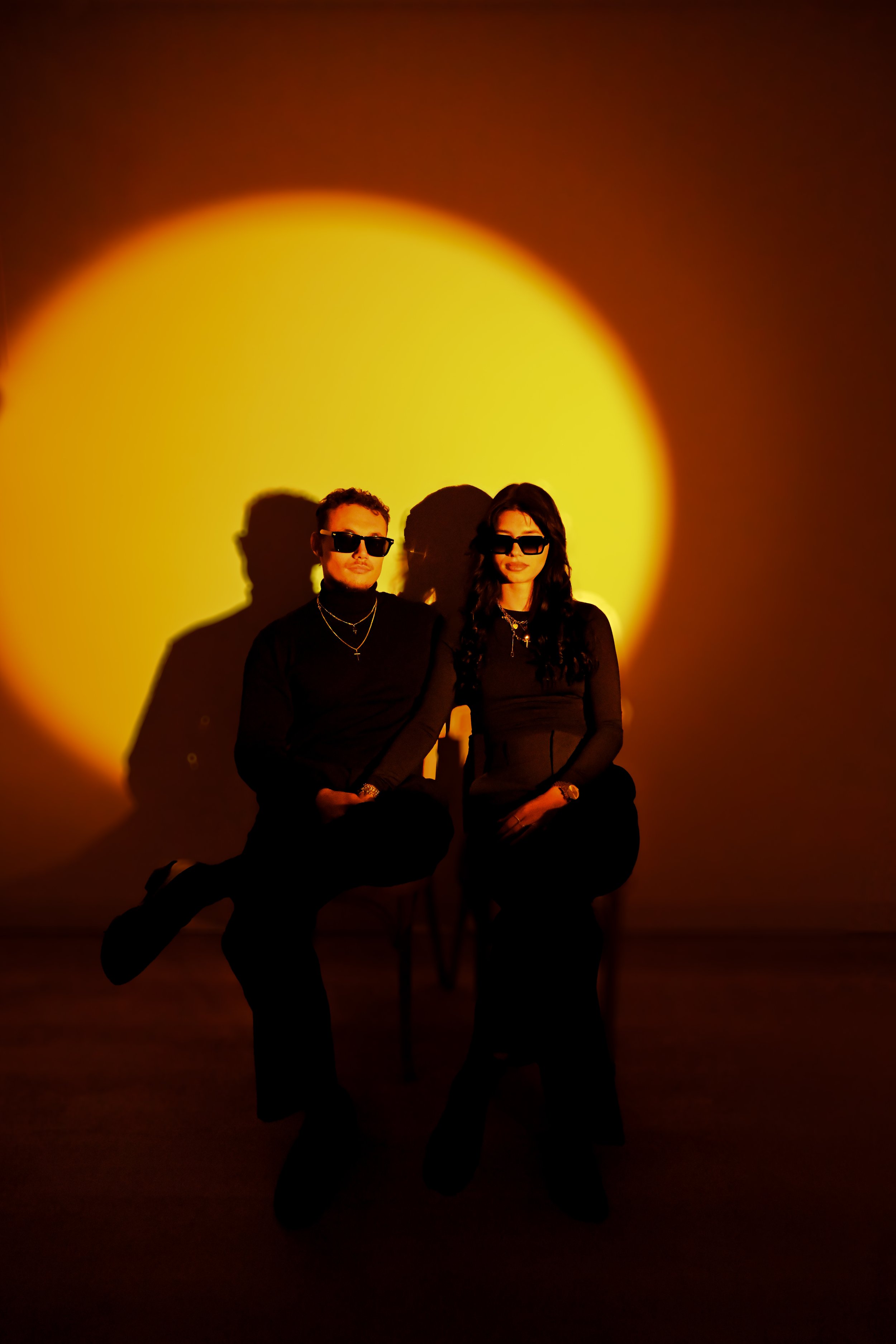 Deux personnes assises devant un mur éclairé par un cercle de lumière jaune, portant des lunettes de soleil noires et des vêtements noirs, créant de longues ombres derrière elles.