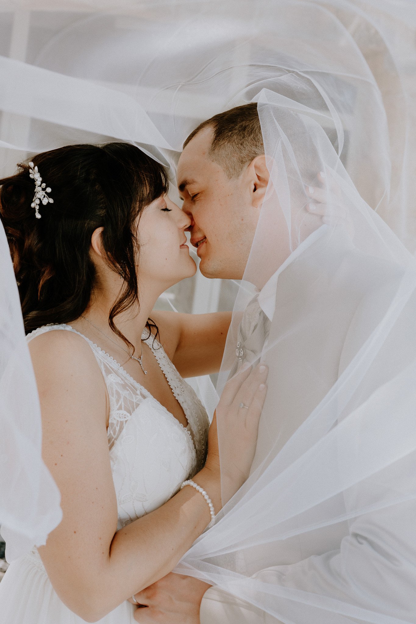 Un couple de mariés se tient à proximité, avec leur nez et leur front qui se touchent, sous une voile de mariage blanche.