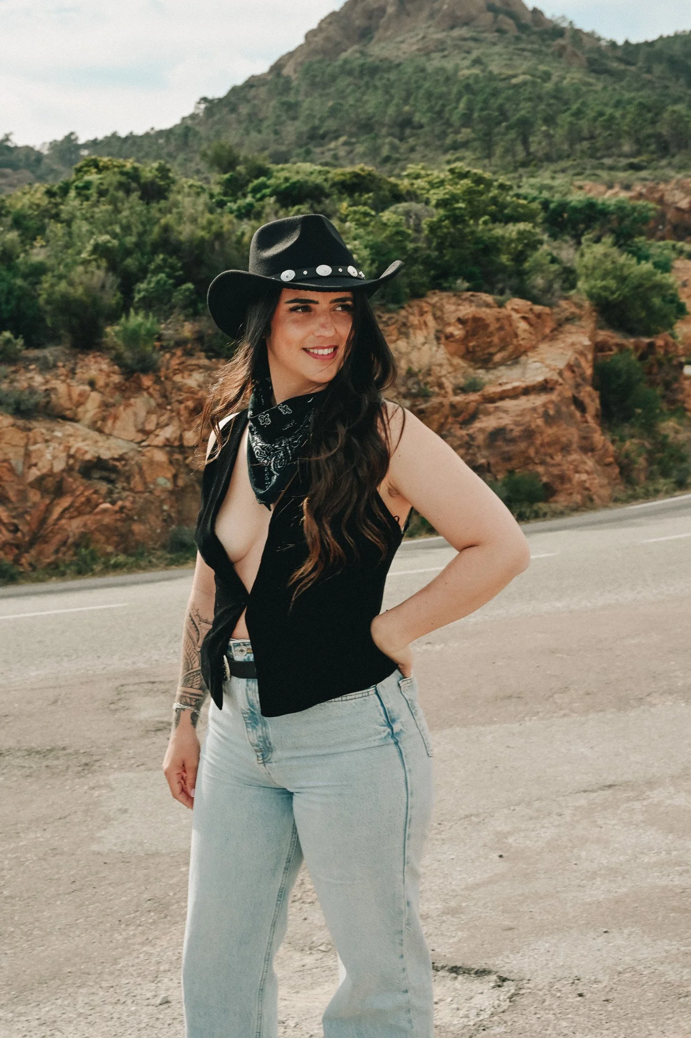 Une femme portant un chapeau cowboy noir et une bandana noire autour du cou, posant à côté d'une route dans un paysage désertique avec des montagnes et des végétation verte en arrière-plan.