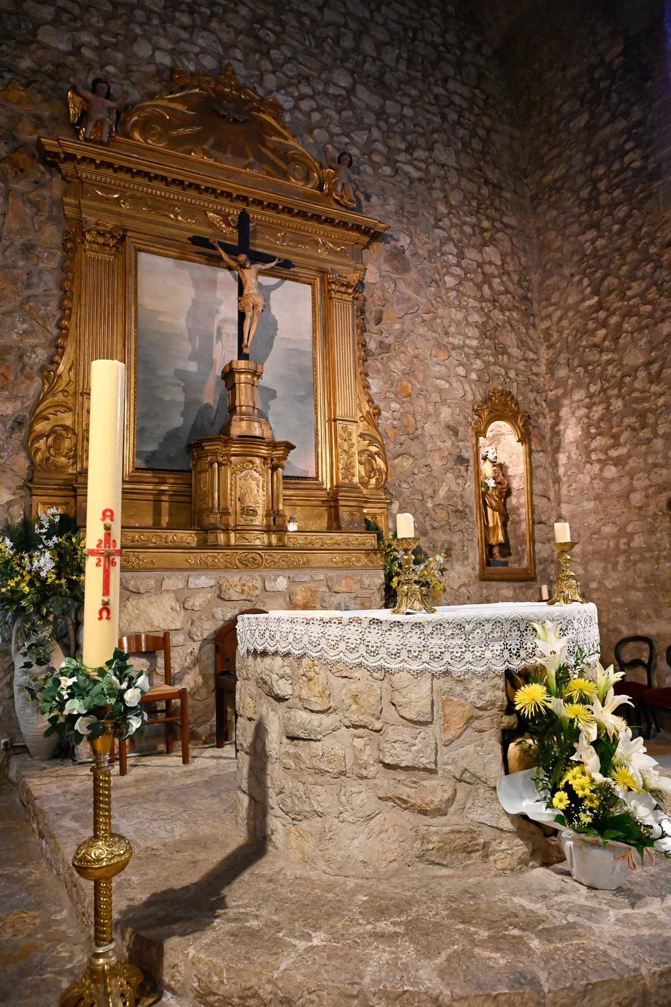 Une petite chapelle ou un lieu de culte en pierre avec un autel en pierre, décoré de fleurs, de bougies et de statues religieuses. Un crucifix en bois orné de dorure est en avant de l'autel, encadré d'une très belle structure en bois et ornementée.