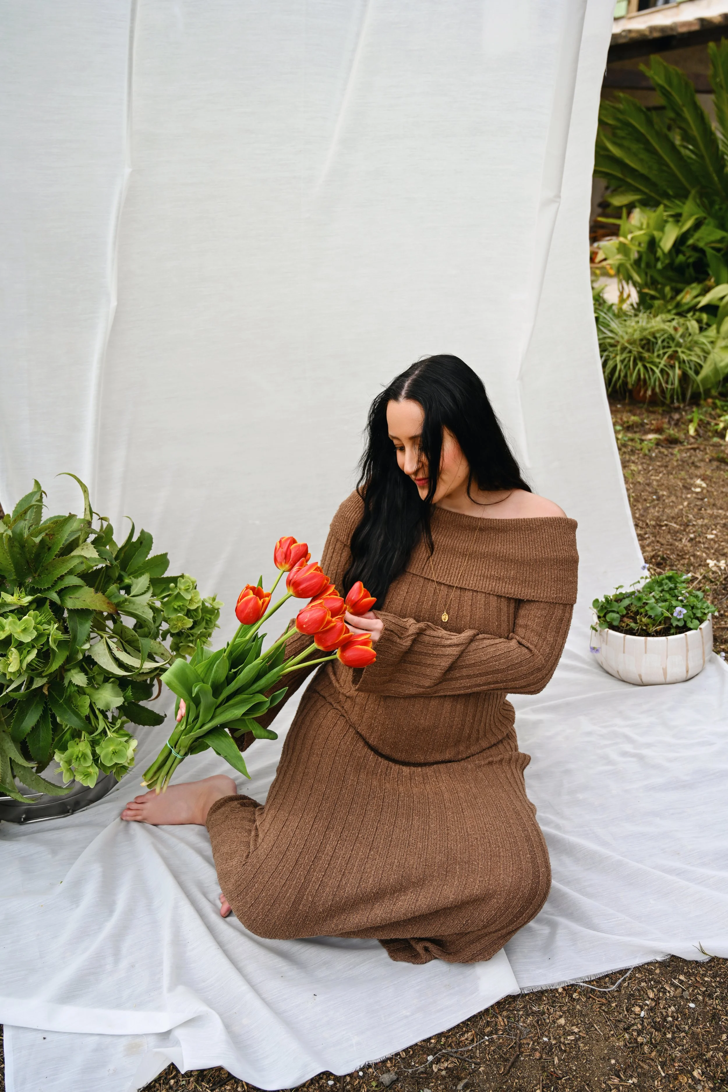 Une femme vêtue d'une robe marron en tricot est assise sur un drap blanc, en train de cueillir des tulipes rouges. Elle est entourée de plantes vertes en pots, dans un jardin.