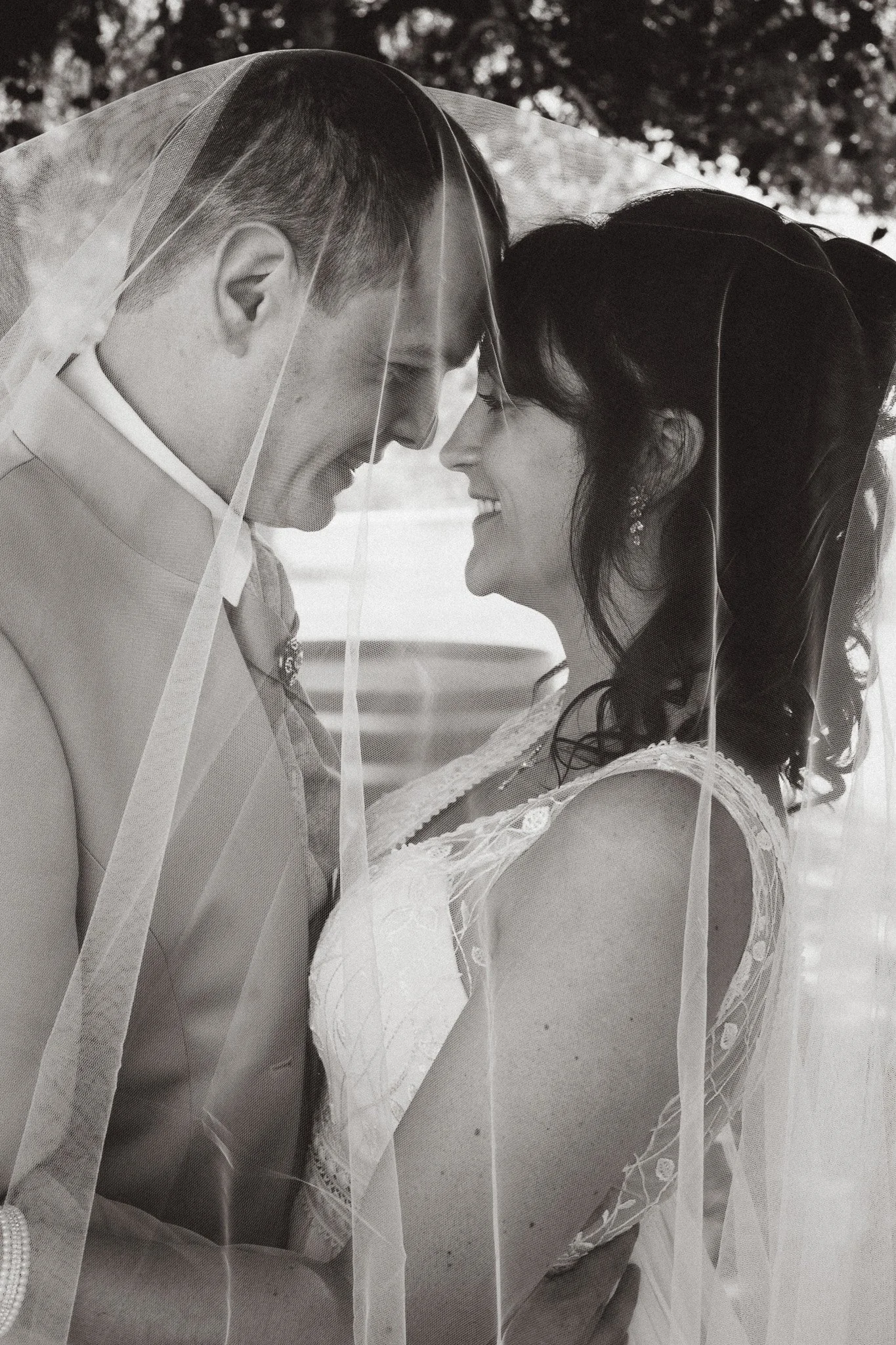 Un couple lors de leur mariage, portant une robe de mariée et un costume, partageant un moment intime sous un voile de mariée, en noir et blanc.