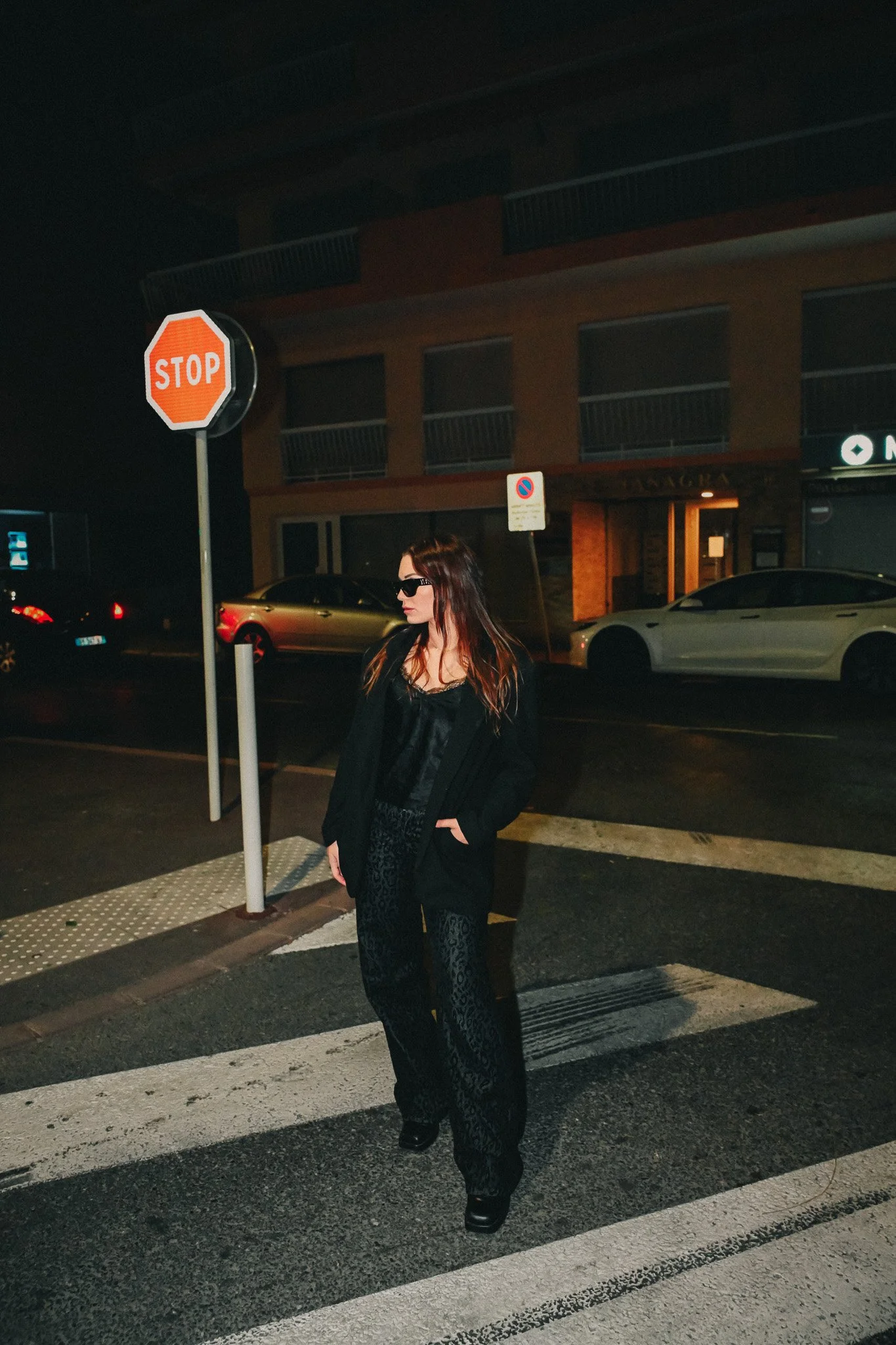 Une femme avec des cheveux longs portant des lunettes de soleil, vêtue en noir, se tenant sur un passage pour piétons la nuit. En arrière-plan, une voiture blanche, une voiture noire, un bâtiment avec des fenêtres et un panneau 'STOP' visible.