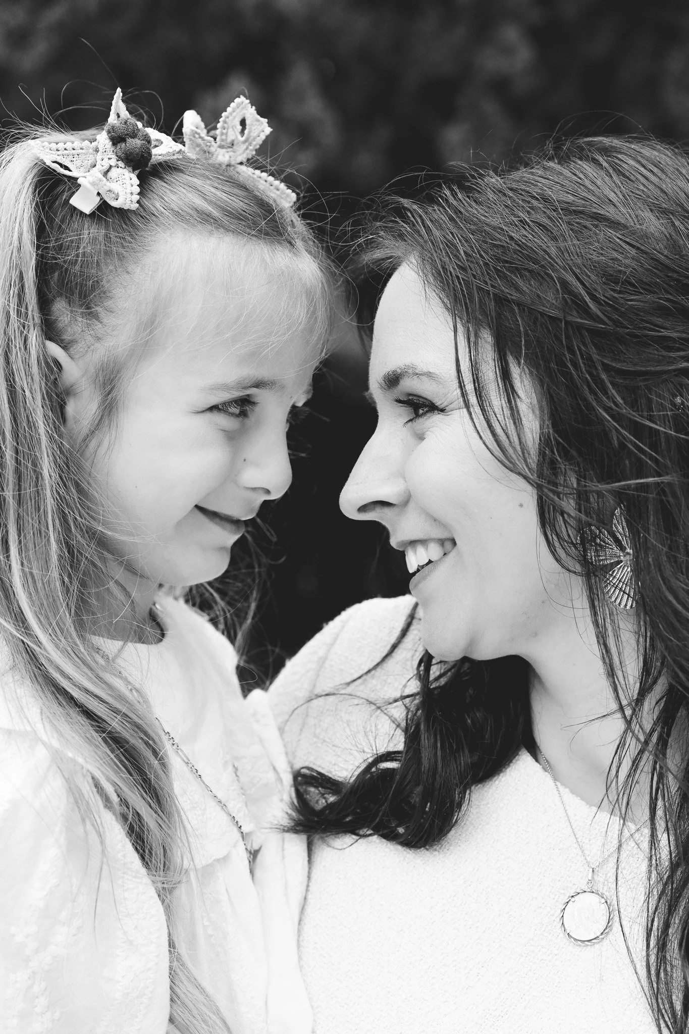 Une femme et une jeune fille, face à face, sourient et se regardent, partageant un moment d'affection. La femme a de longs cheveux foncés et porte un col roulé clair, la jeune fille a des cheveux longs et clairs avec un bandeau en dentelle avec une d
