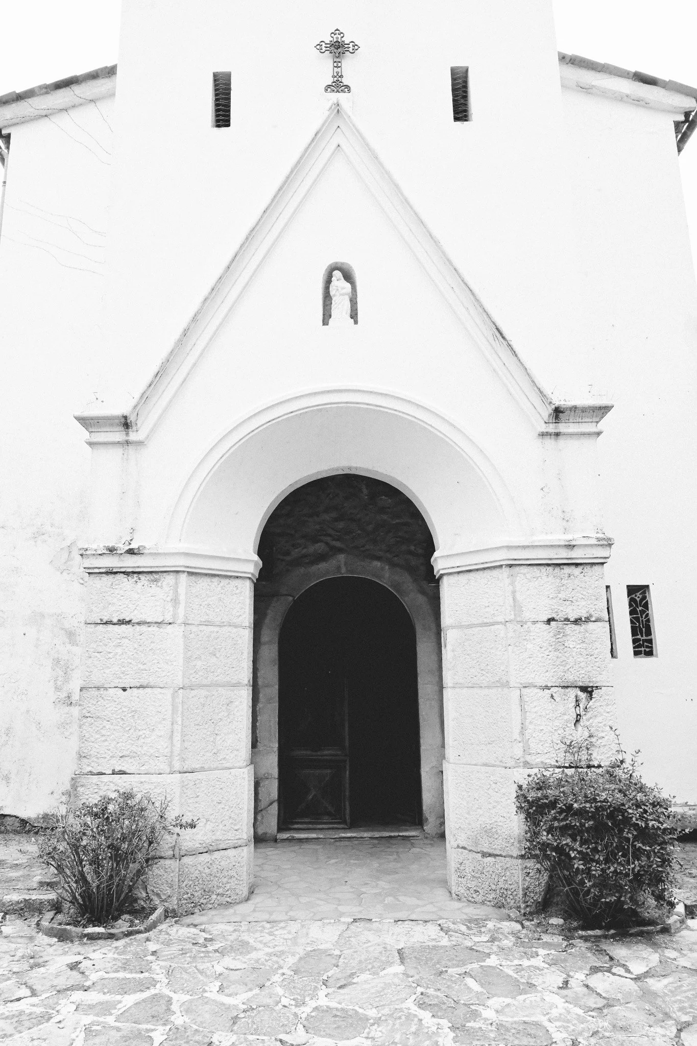 Façade d'une petite église blanche avec un arc d'entrée en pierre, une porte en bois, des arêtes de toit en tuiles, et des fenêtres verticales. Il y a des buissons de chaque côté de la porte en pierre.