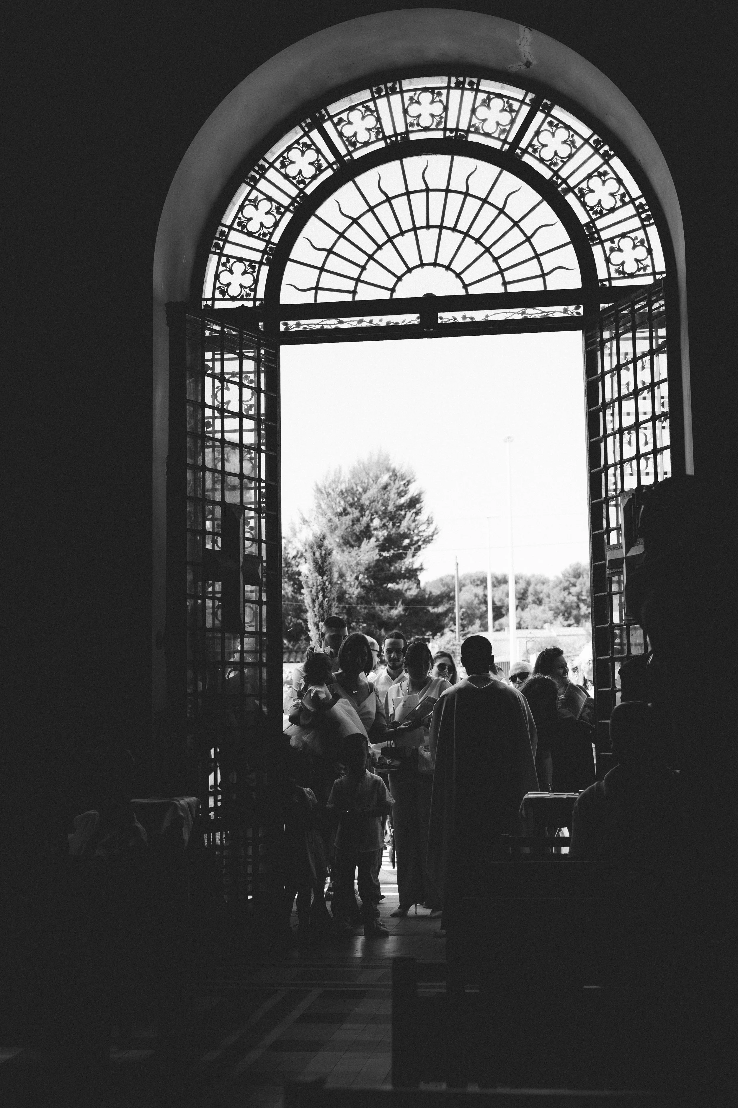 Une scène prise depuis l'intérieur d'un bâtiment avec une grande porte ou fenêtre en métal ouvrant sur l'extérieur. De nombreuses personnes se rassemblent, certains discutant, d'autres tenant des documents ou engagés dans une activité collective. La 