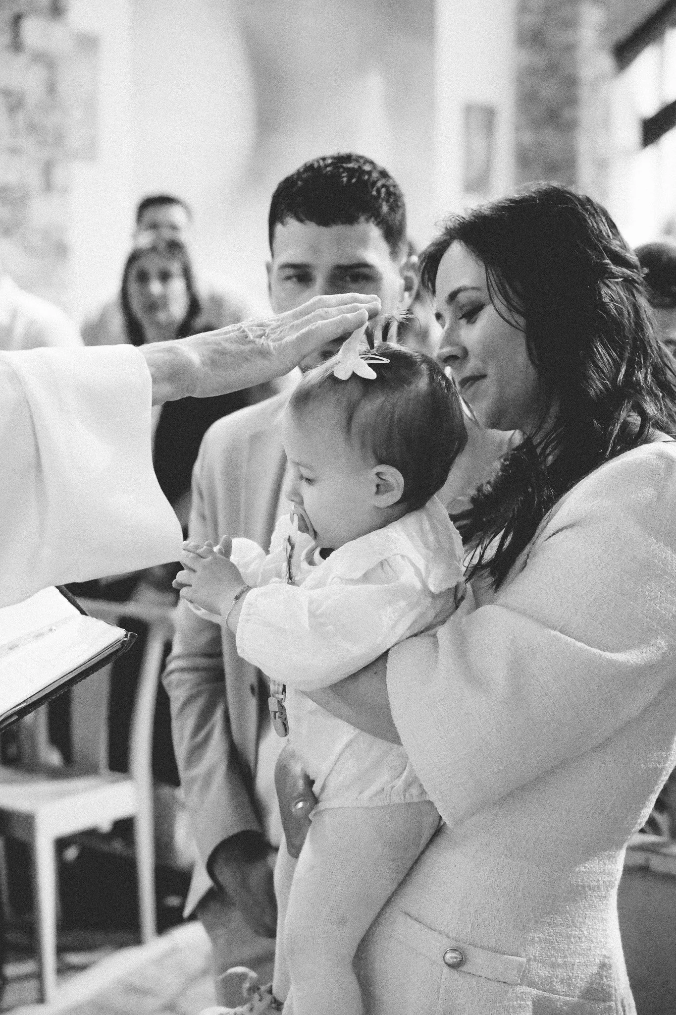 Une femme reçoit une bénédiction avec un geste de la main d'un religieux lors d'une cérémonie, un enfant est dans ses bras, entourée de plusieurs personnes dans une ambiance religieuse.