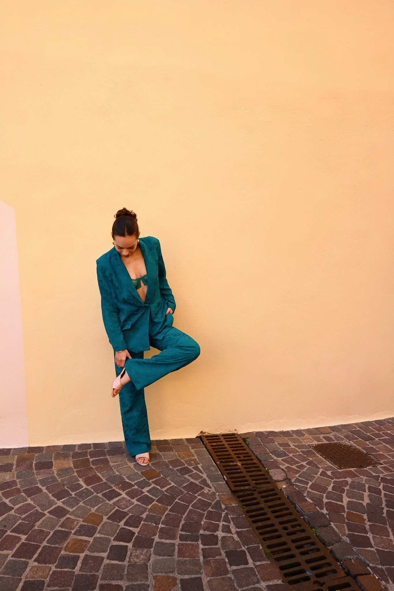 Femme en tailleur en velours vert foncé, posant contre un mur beige, regardant vers le bas, sur un sol en pavés.