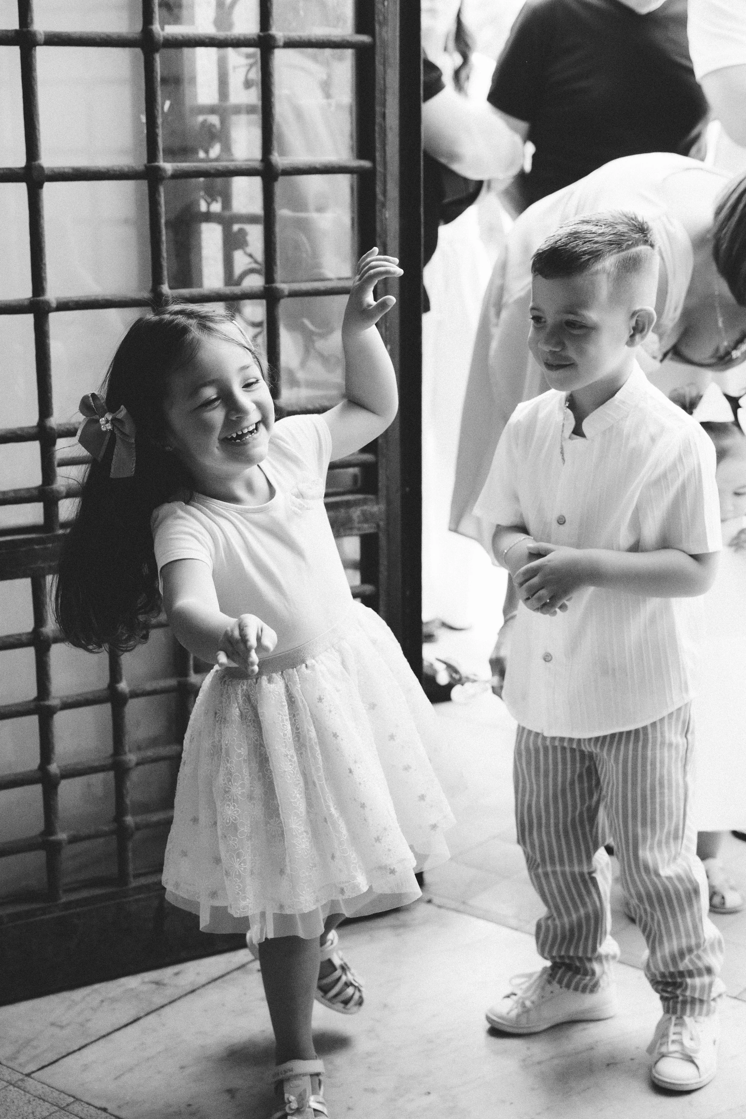 Deux enfants, une fille et un garçon, sourient et dansent dans une pièce. La fille porte une robe blanche et des chaussures à sandales, le garçon porte un pantalon à rayures et une chemise. Des adultes sont flous en arrière-plan.