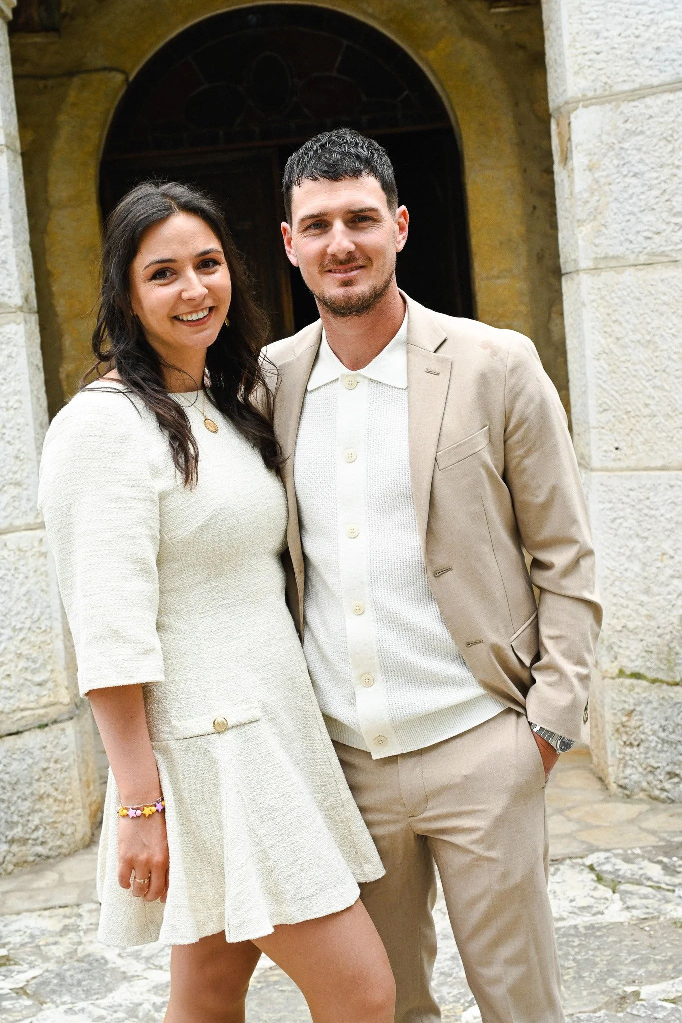 Un homme et une femme souriants posent devant un bâtiment en pierre d'aspect ancien, vêtus de vêtements décontractés élégants, dans une ambiance en plein air.