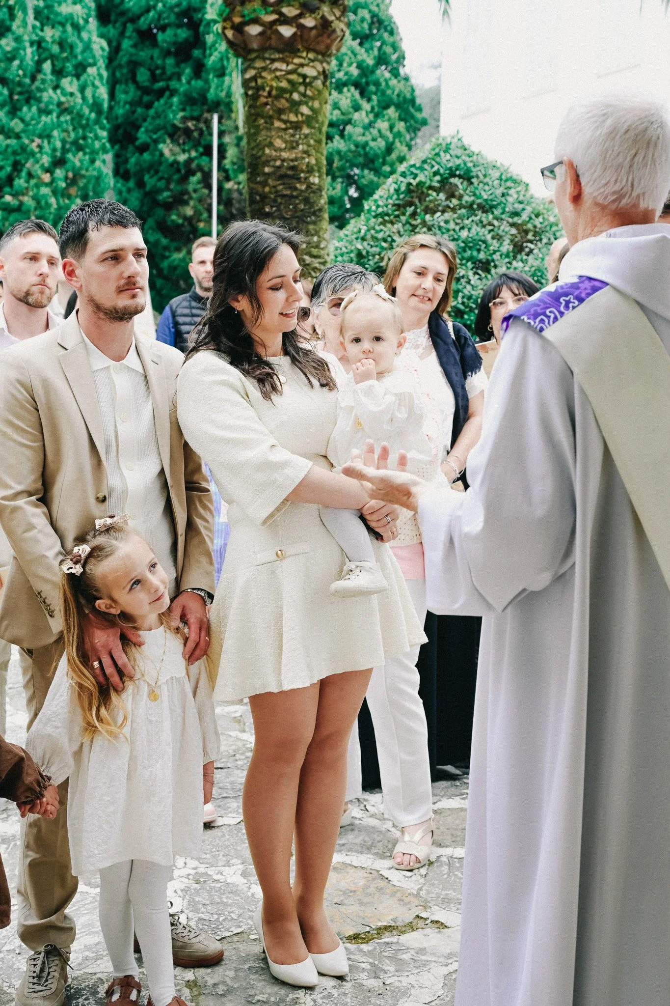 Une cérémonie religieuse avec plusieurs personnes, dont des enfants et un prêtre, en extérieur, avec des arbres en arrière-plan.