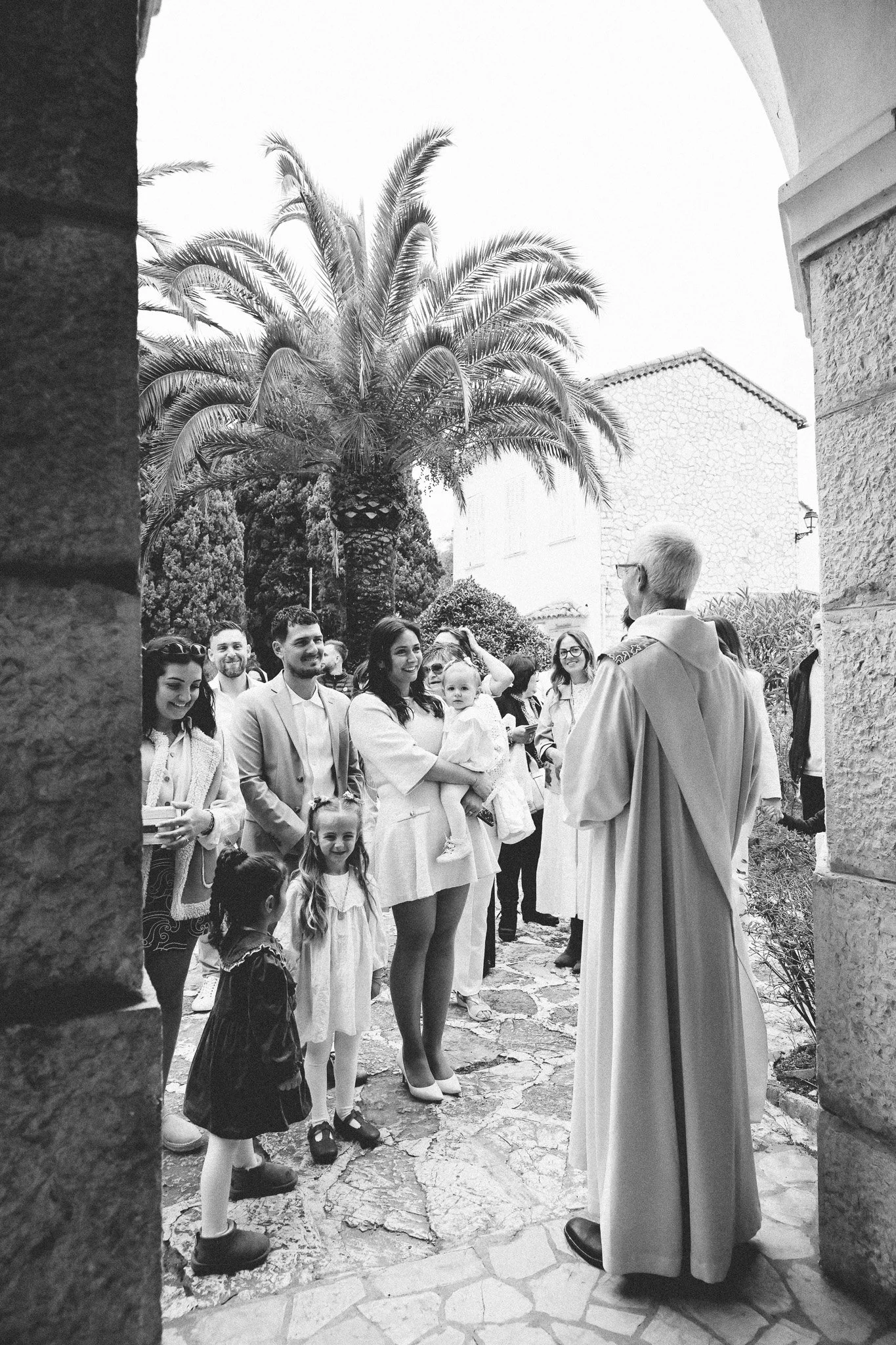 Groupe de personnes, enfants et adultes, réunies à l'extérieur, probablement lors d'une cérémonie ou d'un événement religieux, avec un prêtre ou un pasteur au centre, sous un palmier, en tenue formelle et décontractée.