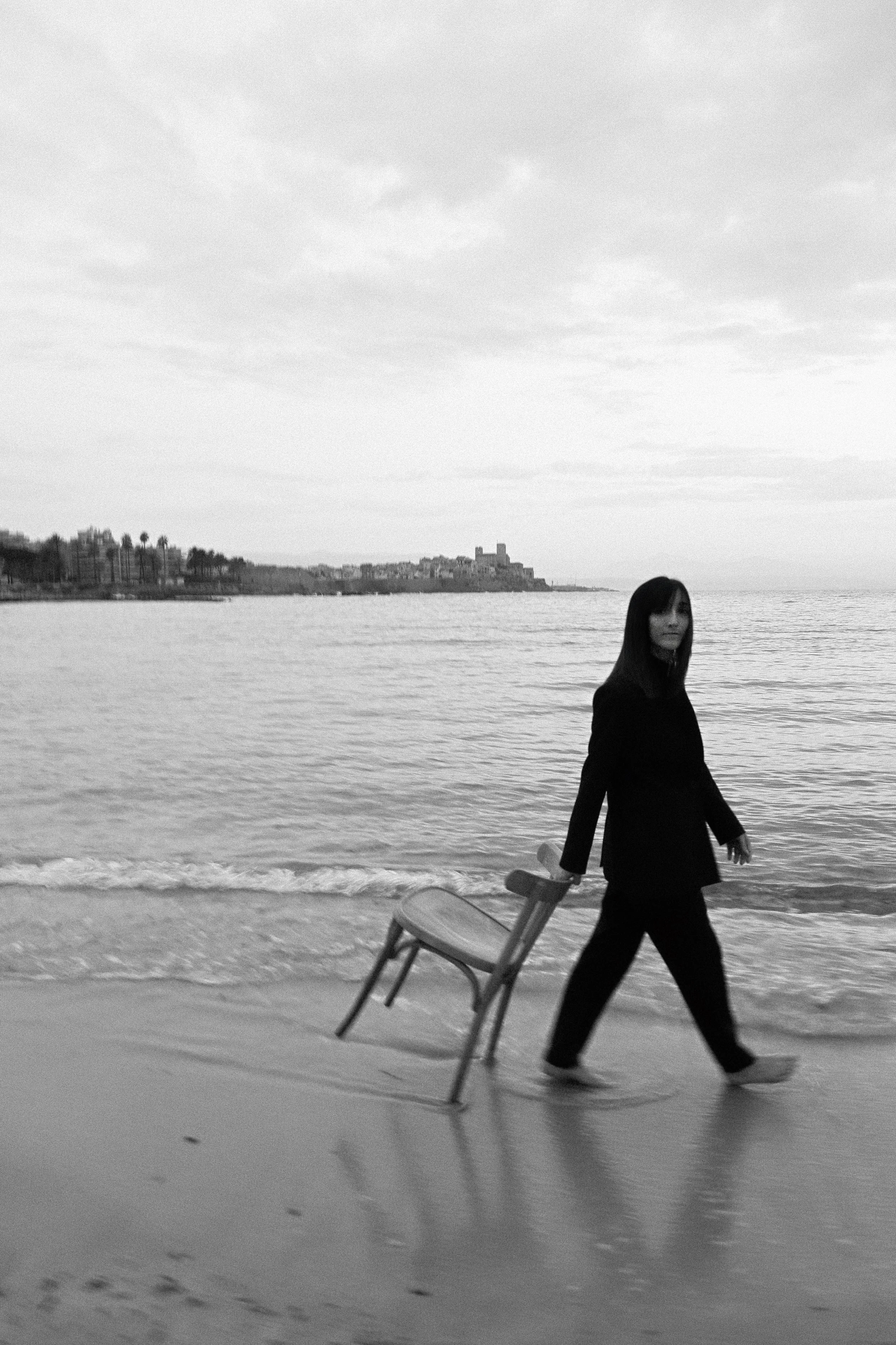 Femme marchant sur la plage avec une chaise pliante à la main, vue en plan inclinée, en noir et blanc, avec des bâtiments en arrière-plan et des nuages dans le ciel.