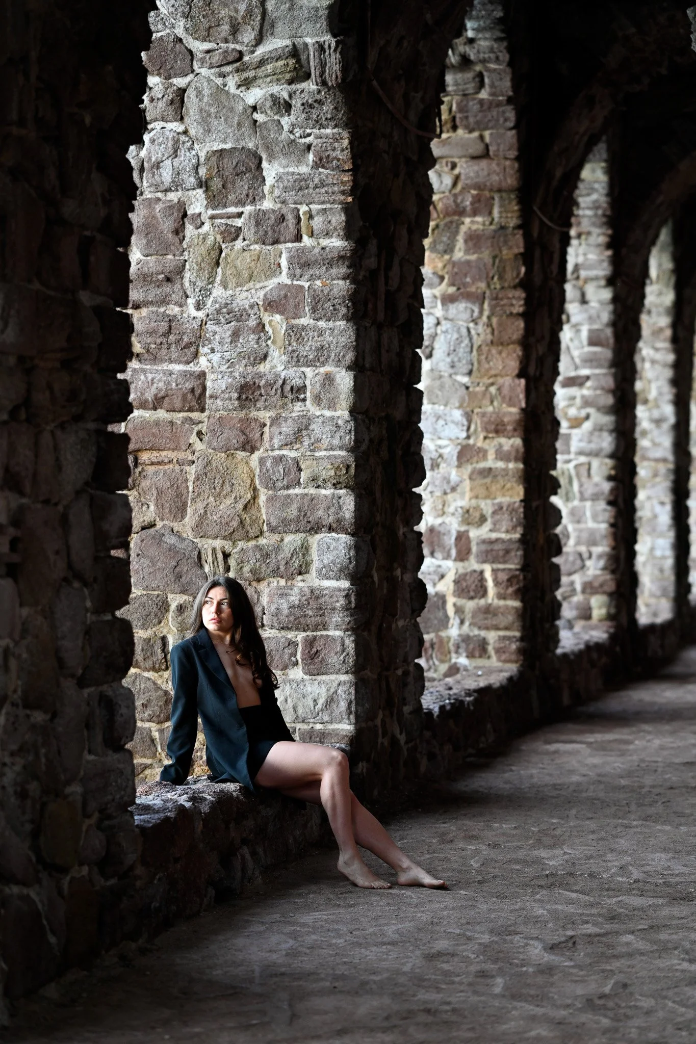 Une femme assise sur un rebord en pierre dans un bâtiment en ruines en pierre, portant une veste noire et une jupe courte, avec des murs en pierre brune et des arches en arrière-plan.