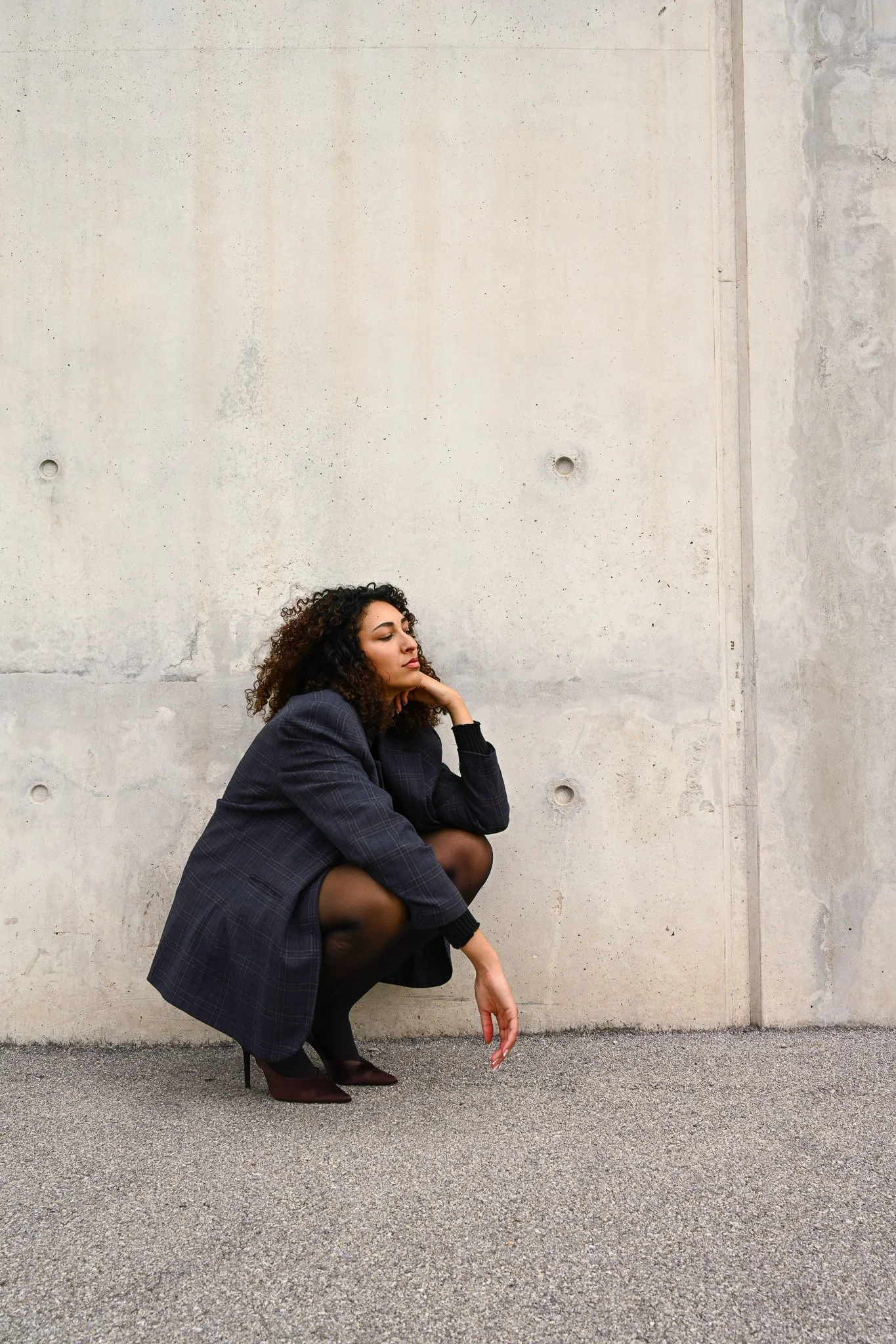 Une femme portant un costume oversize de couleur foncée, en position accroupie contre un mur en béton, avec une main près du visage et portant des talons hauts.