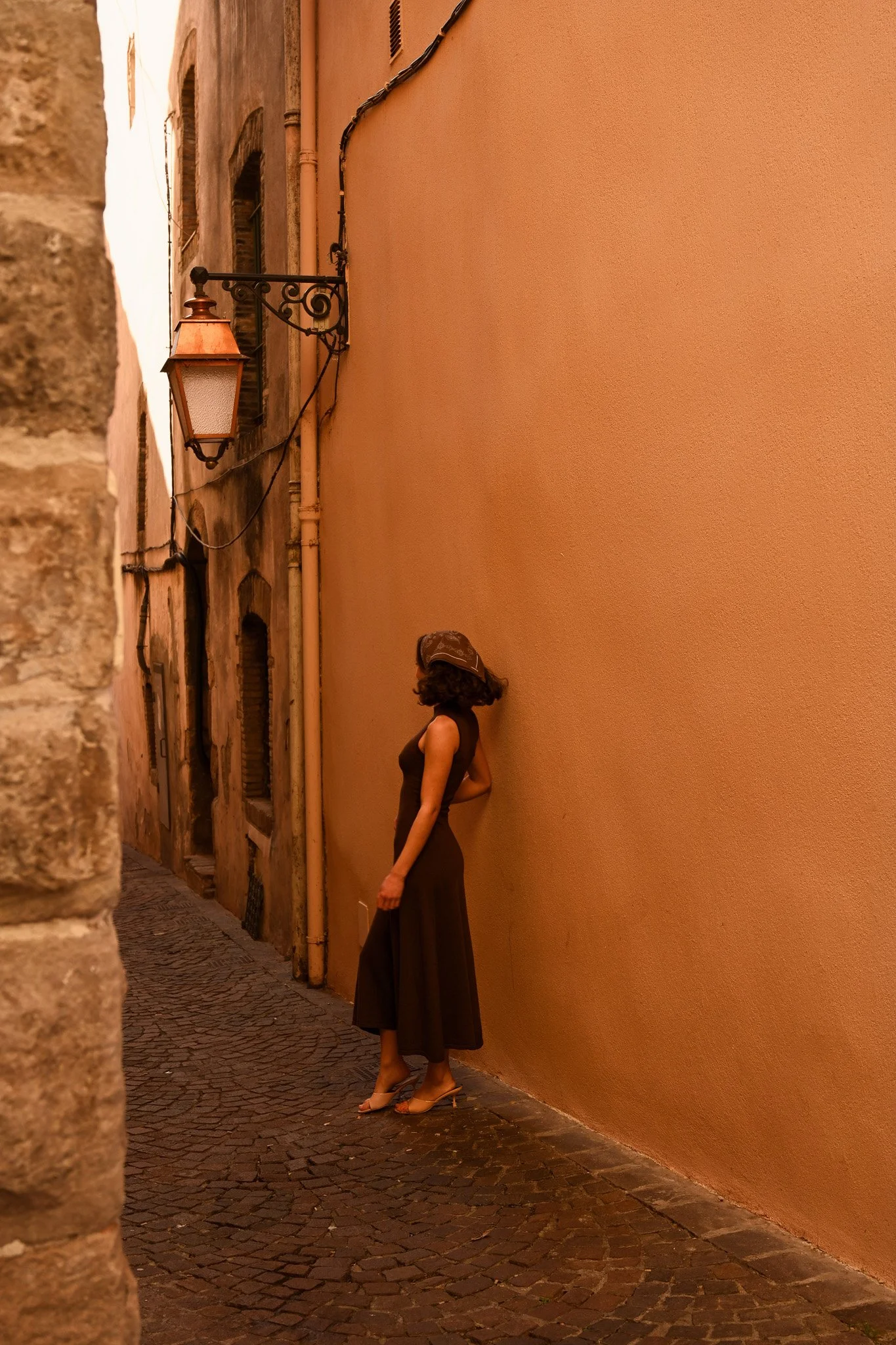 Une femme en robe noire et talons hauts, portant un chapeau, se tient contre un mur beige dans une ruelle pavée, entourée de bâtiments urbains au coucher de soleil.
