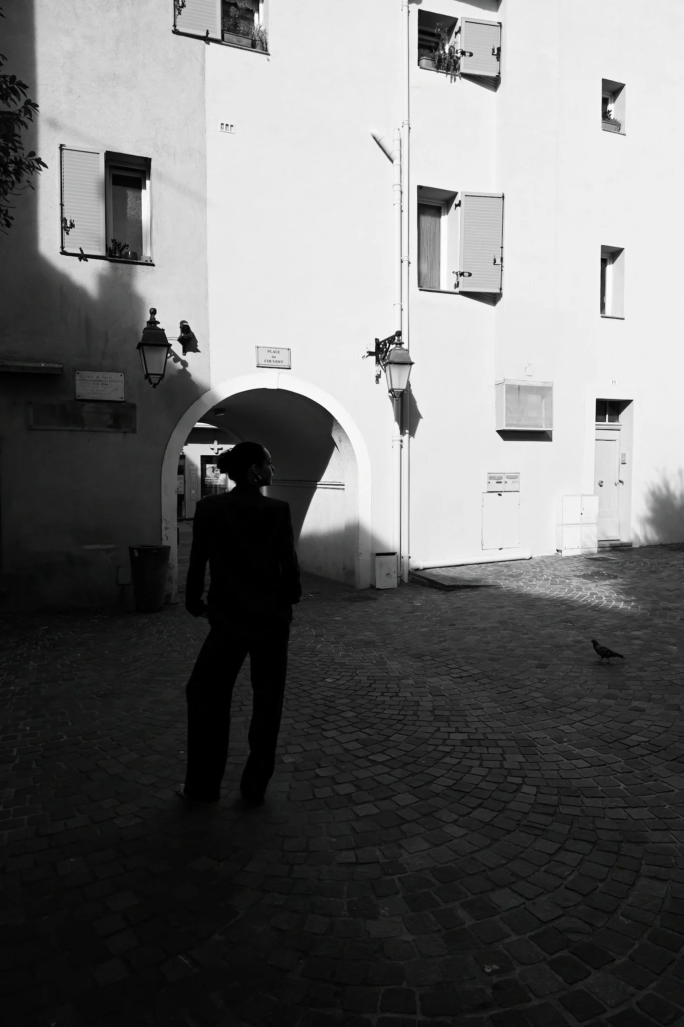Une femme silhouette en noir se tenant dans une rue pavée, en face d'un bâtiment blanc avec plusieurs fenêtres, une arche d'entrée, et des lampadaires. L'image est en noir et blanc.