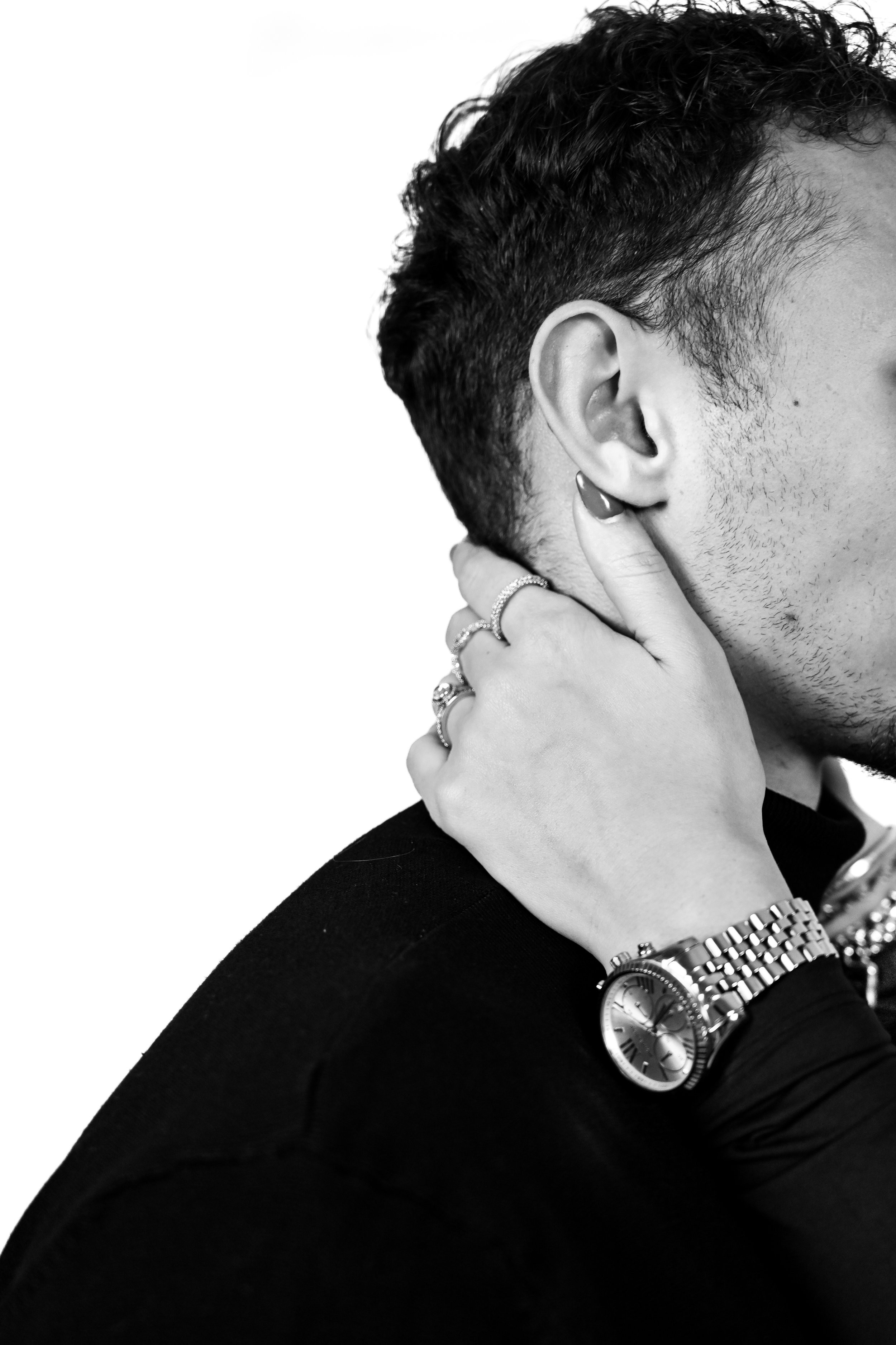 Close-up d'une personne avec des cheveux foncés, portant une montre, plusieurs bracelets, et des bagues, posant avec la main près de l'oreille, en noir et blanc.