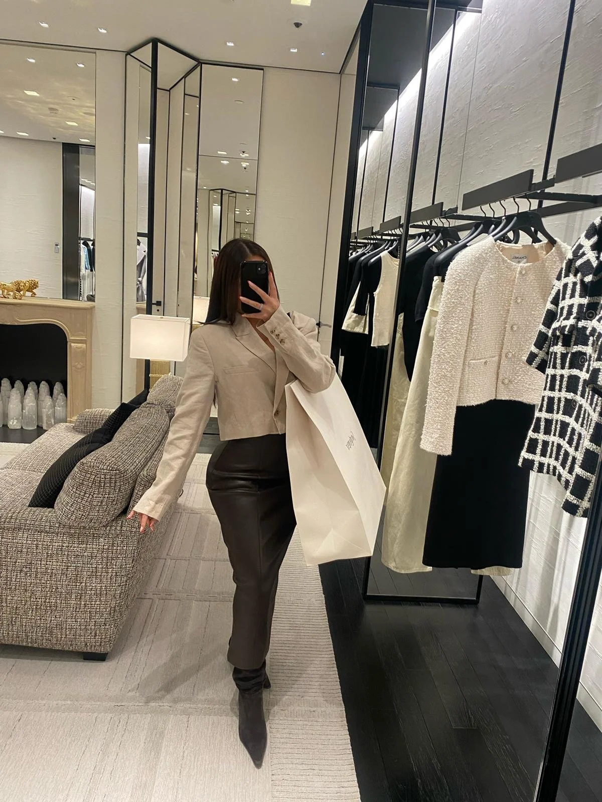 Une femme dans une boutique de vêtements prend une selfie devant un miroir. Elle porte un blazer beige, une jupe en cuir foncé, et des bottines noires. La boutique présente des vêtements noir et blanc suspendus, avec des murs en blanc et des miroirs grands et rectangulaires.
