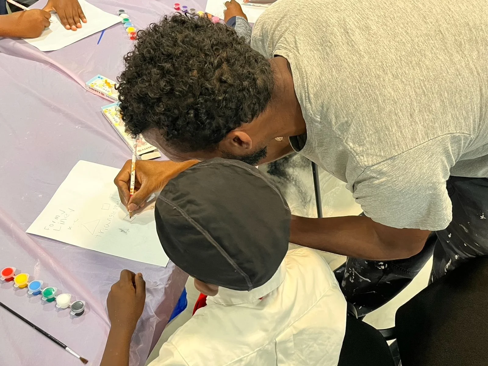 A man with curly hair and a beard is leaning over a table, writing on a large white sheet of paper. A child in a tan shirt and a black cap is sitting at the table, looking at the paper. Paints and other art supplies are on the table, which is covered with a pink tablecloth.