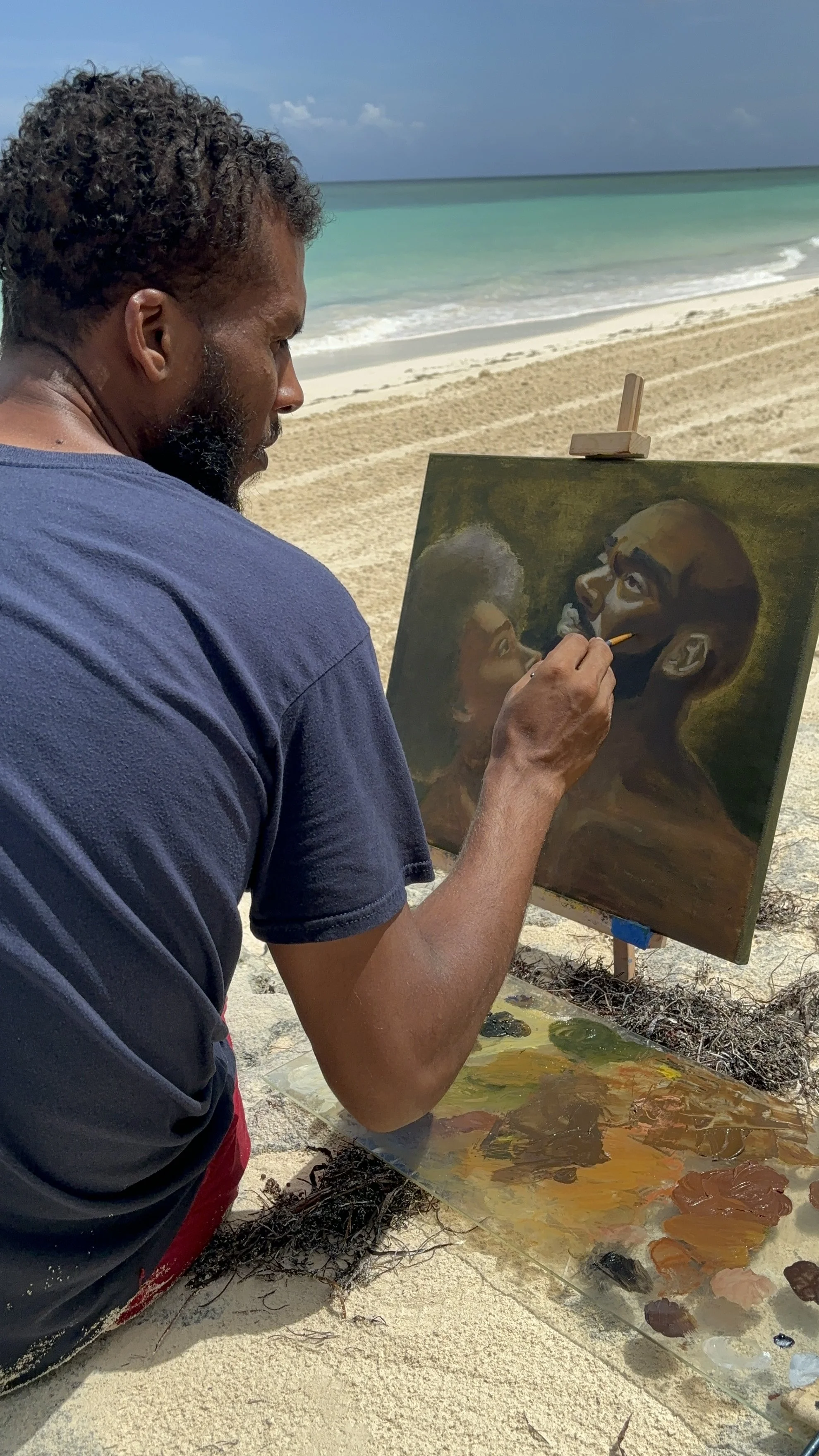 A man painting a portrait of a woman and a man on a canvas set up on the sandy beach, with the ocean and sky in the background.