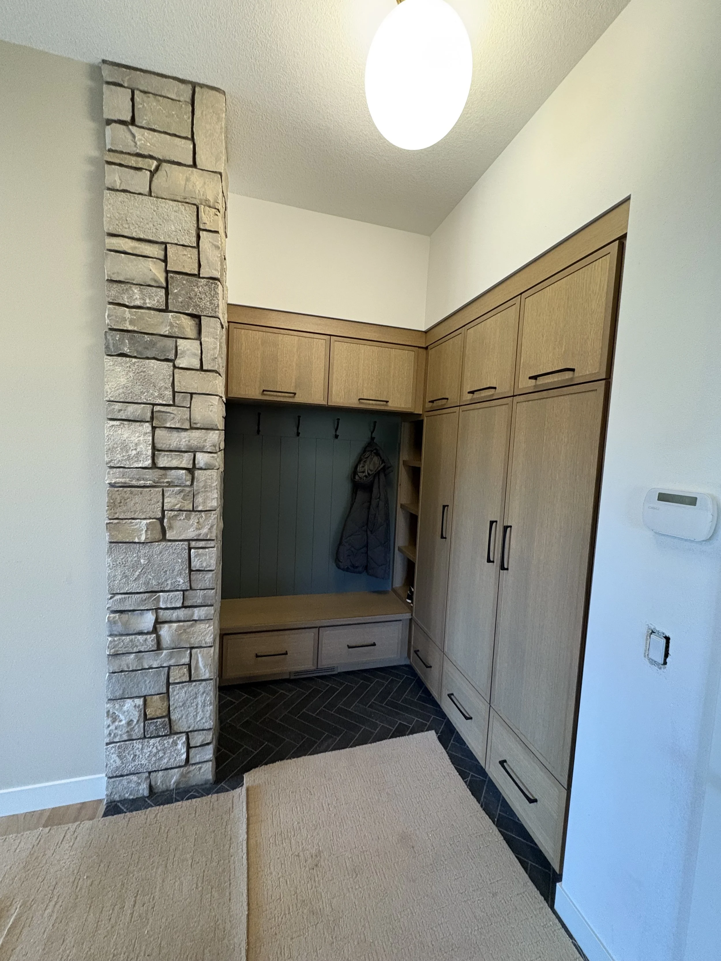 Entryway with wooden cabinets, black bar handles, coat hooks with jacket, stone wall, black herringbone tile floor, beige and cream rugs, white walls, ceiling light, thermostat, and electrical outlet.