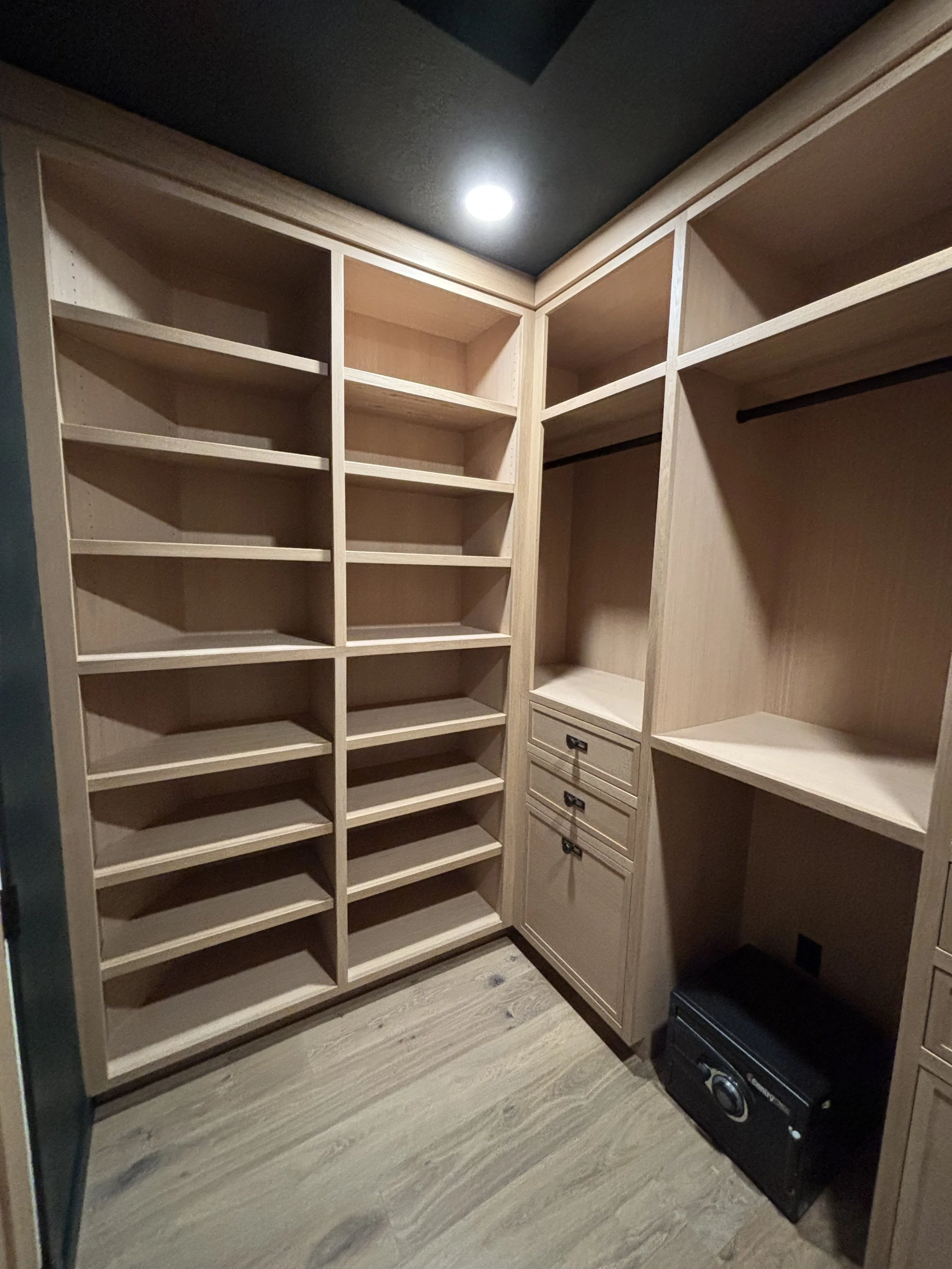 Empty walk-in closet with built-in wooden shelves, drawers, and a hanging rod, with a black safe and small black speaker on the floor.