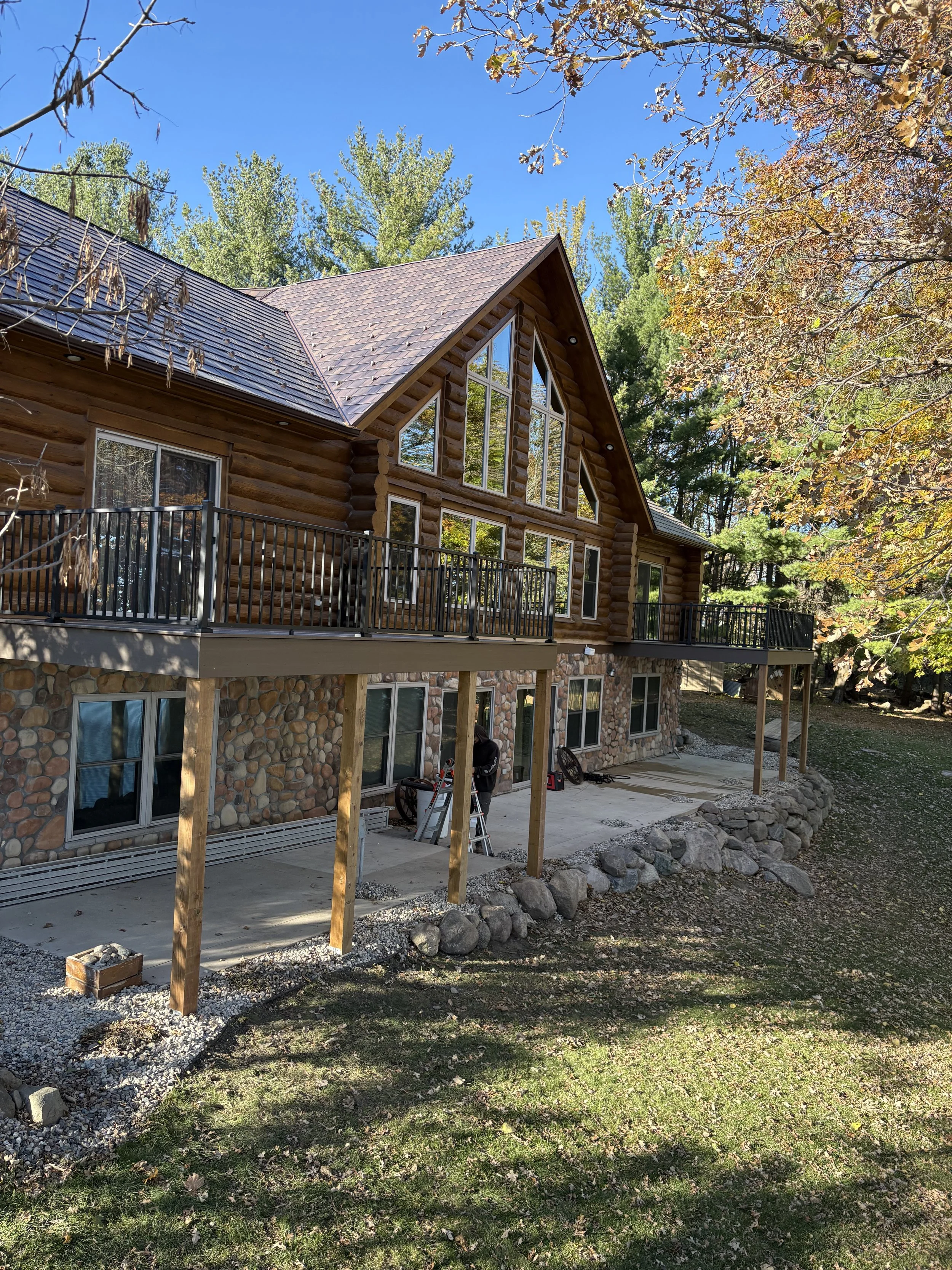 A two-story log cabin with large windows, a sloped roof, and a stone foundation, surrounded by trees with autumn leaves. There is a balcony with a metal railing and some construction work ongoing with wooden support beams and concrete foundation bein