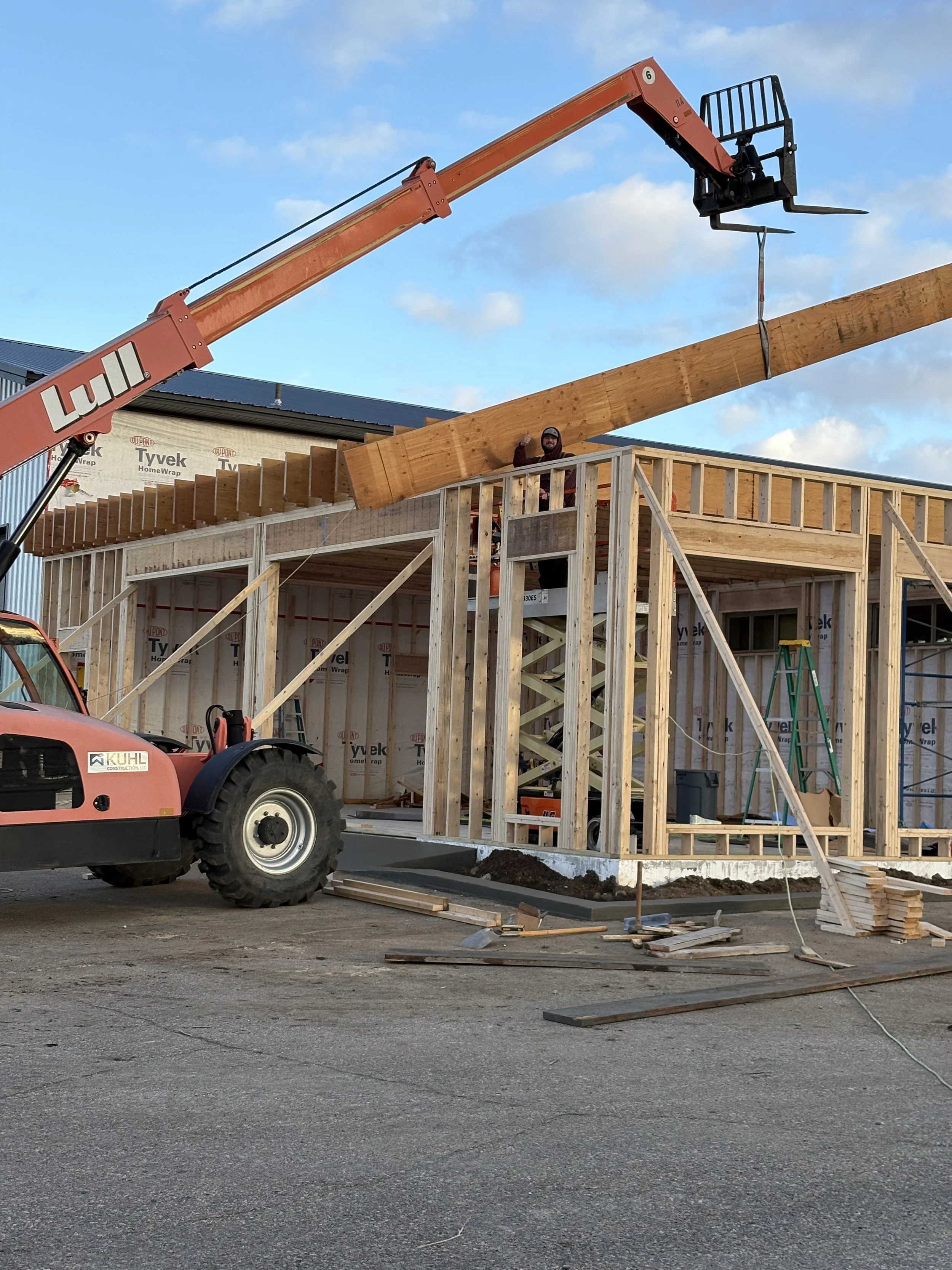 Construction site with a wooden frame house being built, a worker giving a thumbs-up, and a crane lifting a wooden beam.