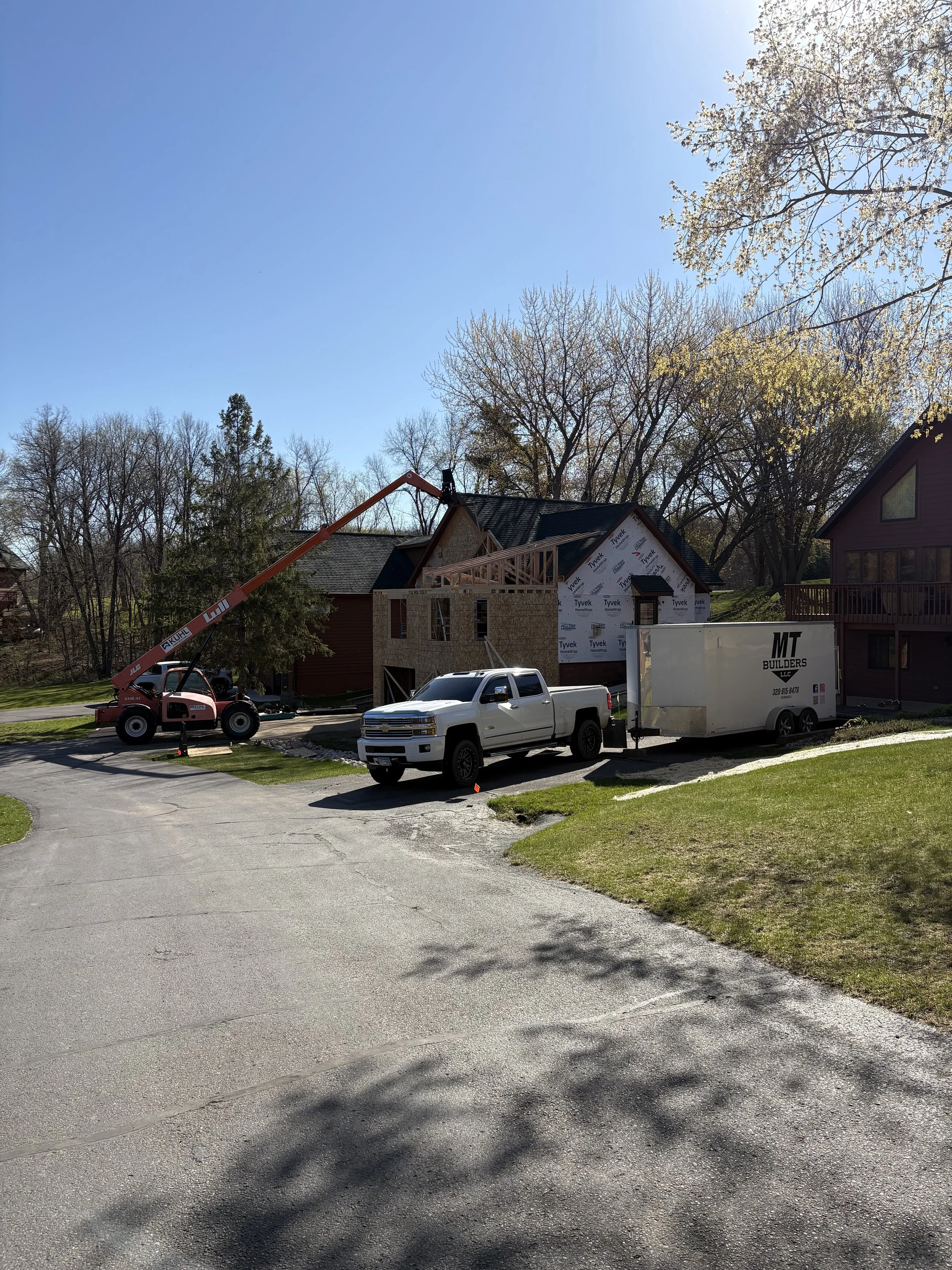 A house under construction with a crane lifting materials, parked white trucks, and trees surrounding the area on a sunny day.