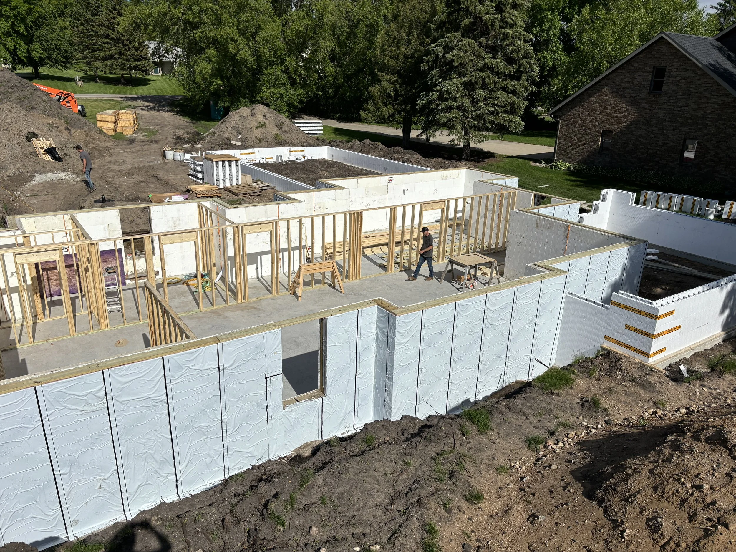 Construction site with foundation and framing in progress, surrounded by dirt and trees, with workers present.