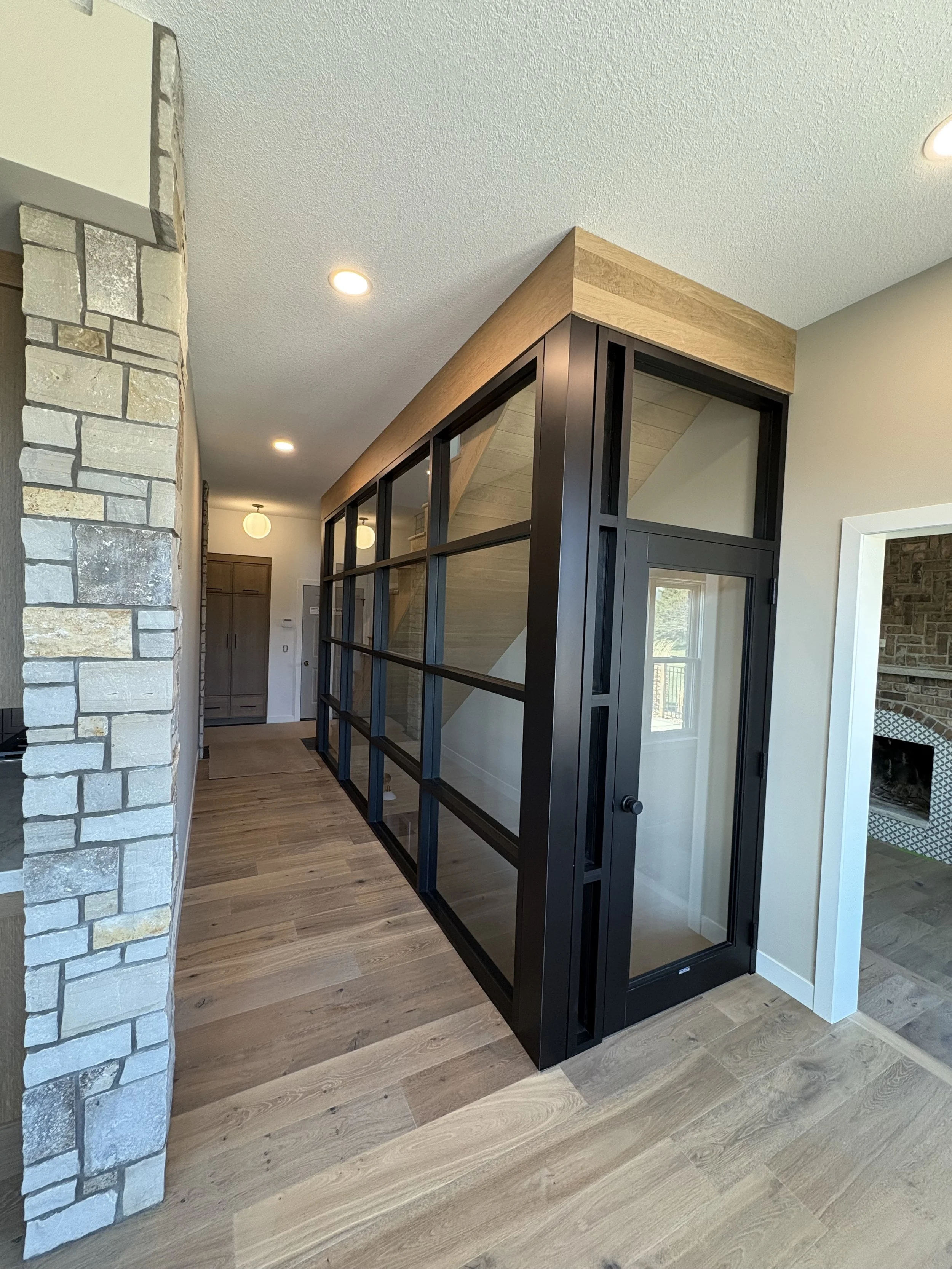 Interior view of a modern house with a glass and black metal staircase, wooden beams, and a stone wall in the background, with hardwood flooring and recessed lighting.