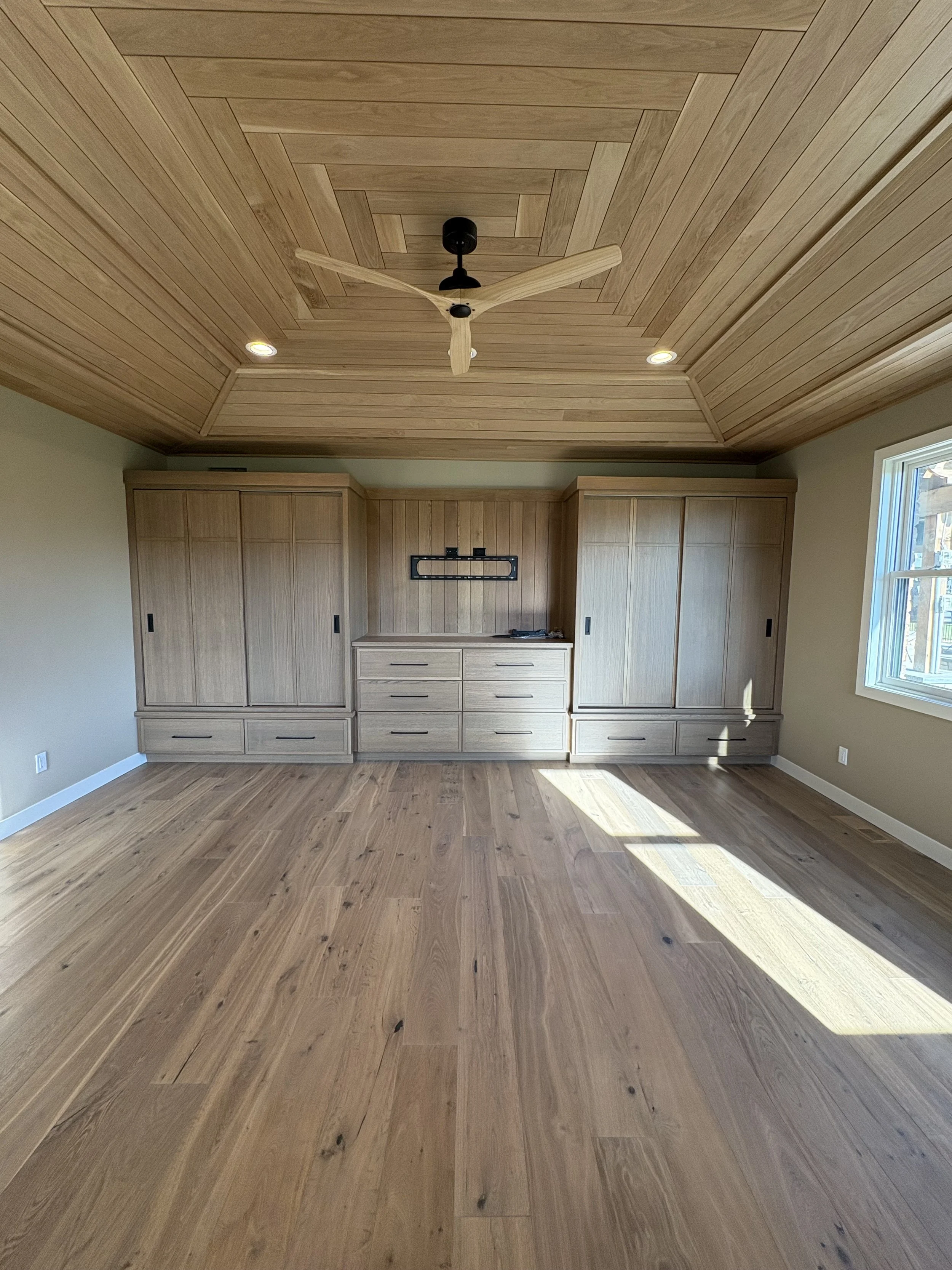 A room with a wooden ceiling, including a ceiling fan, built-in cabinets, and a window.