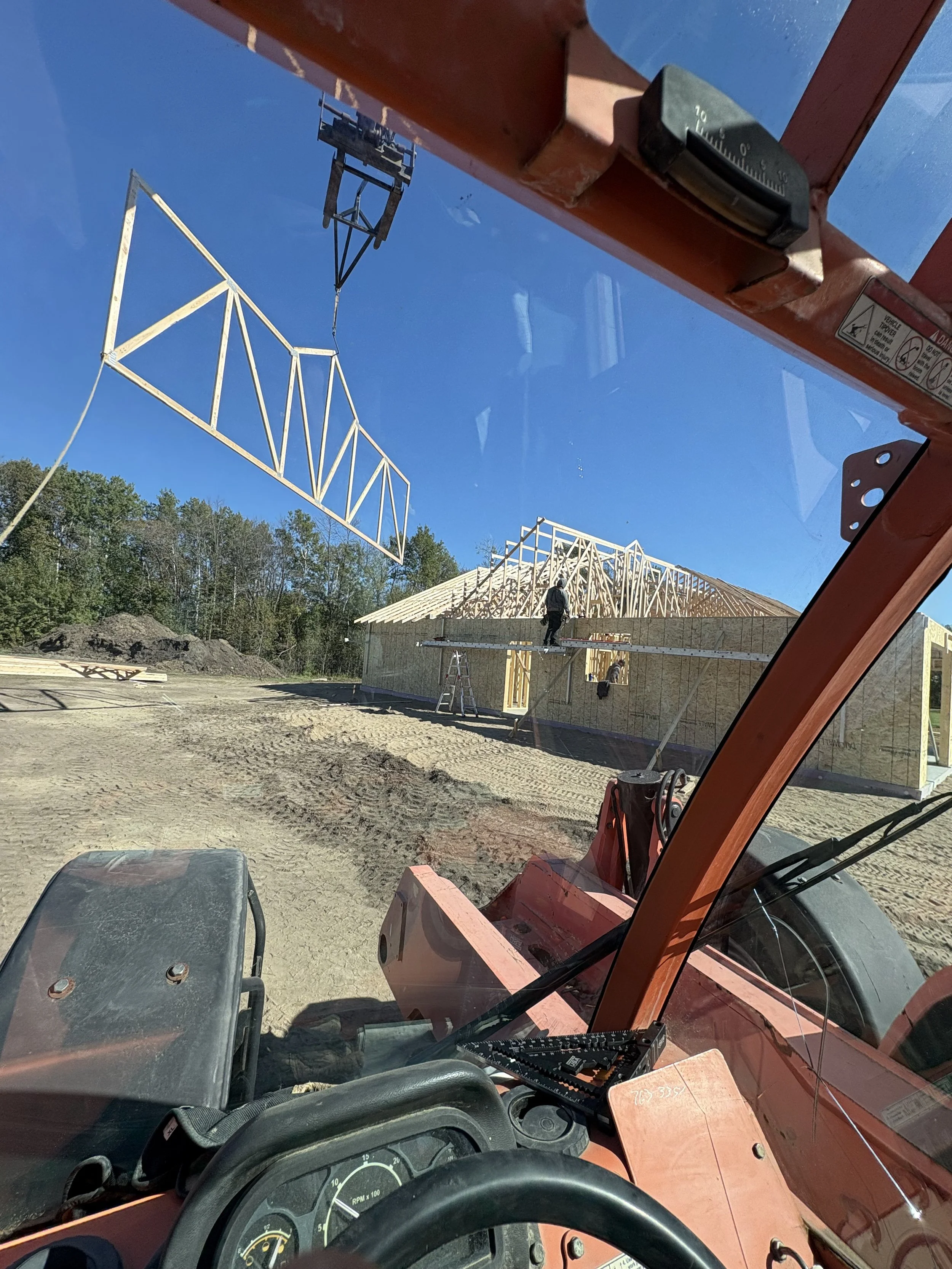 Construction site with wooden house framework, workers, and crane in the background, viewed from inside a construction vehicle.