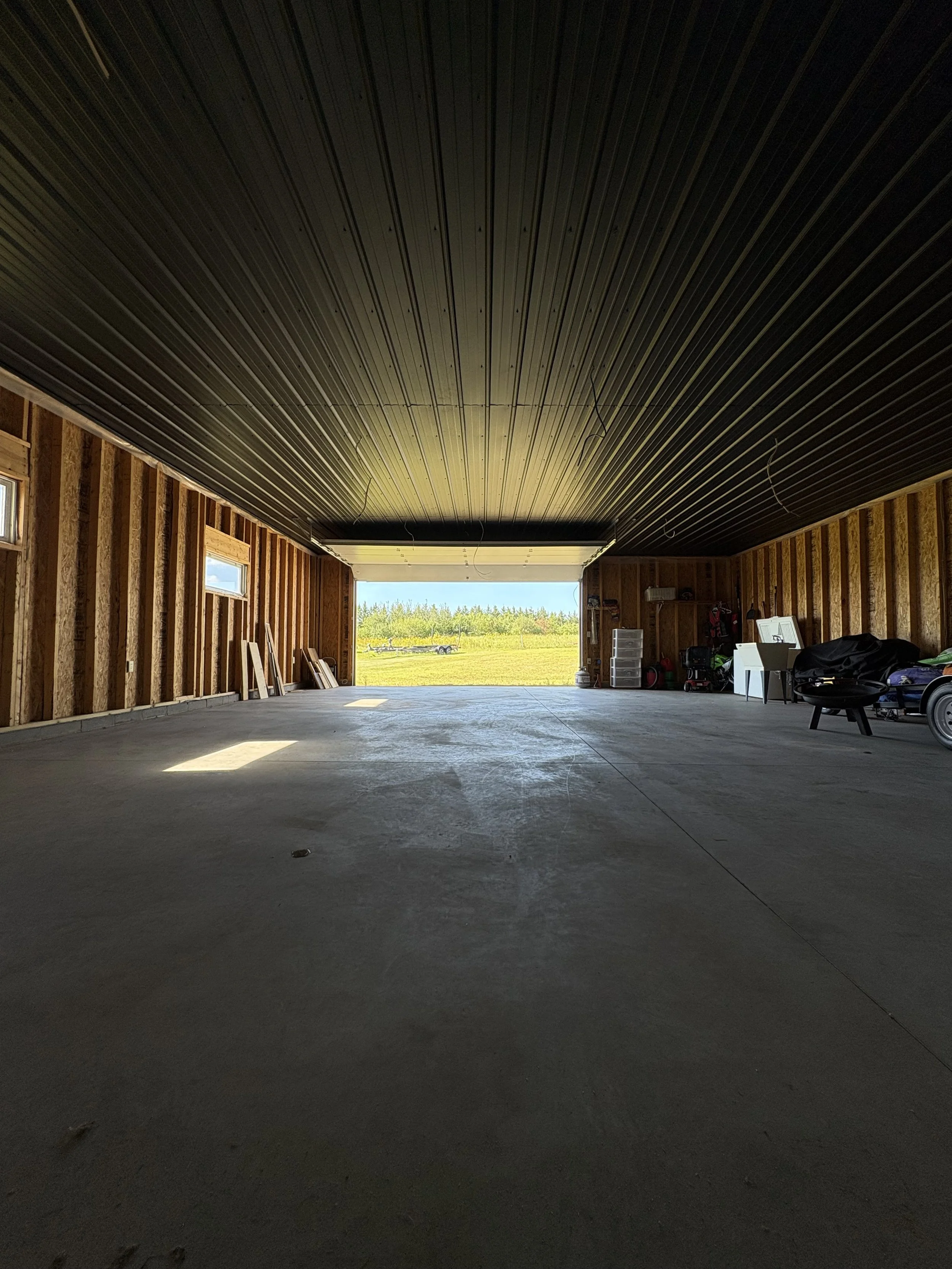 Interior of a partially constructed garage with exposed wooden framing, a concrete floor, and an open garage door showing a view of grass and trees outside.