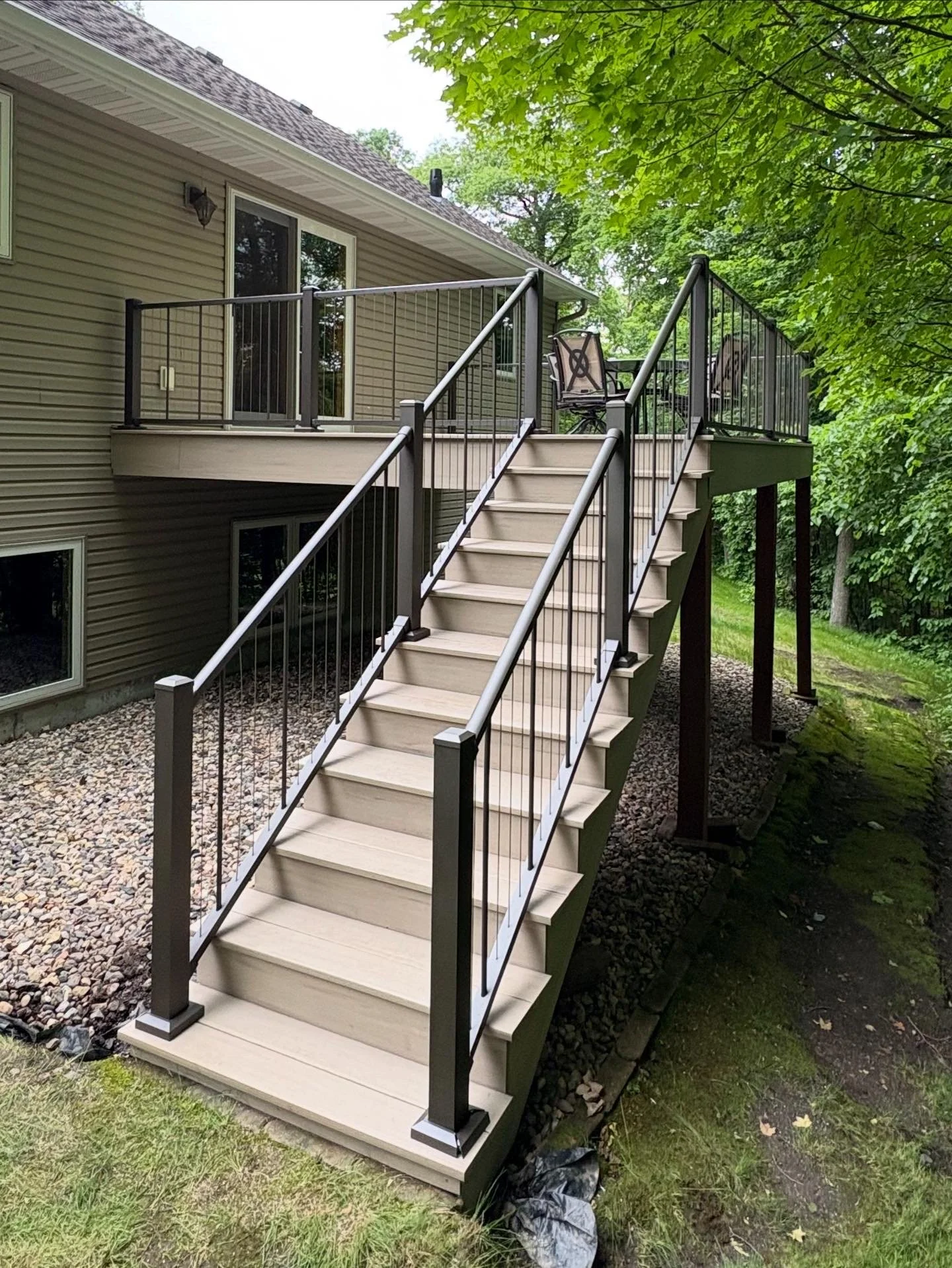 A newly built wooden outdoor deck with metal railings attached to a beige house, stairs leading down to the yard, surrounded by green trees and grass.