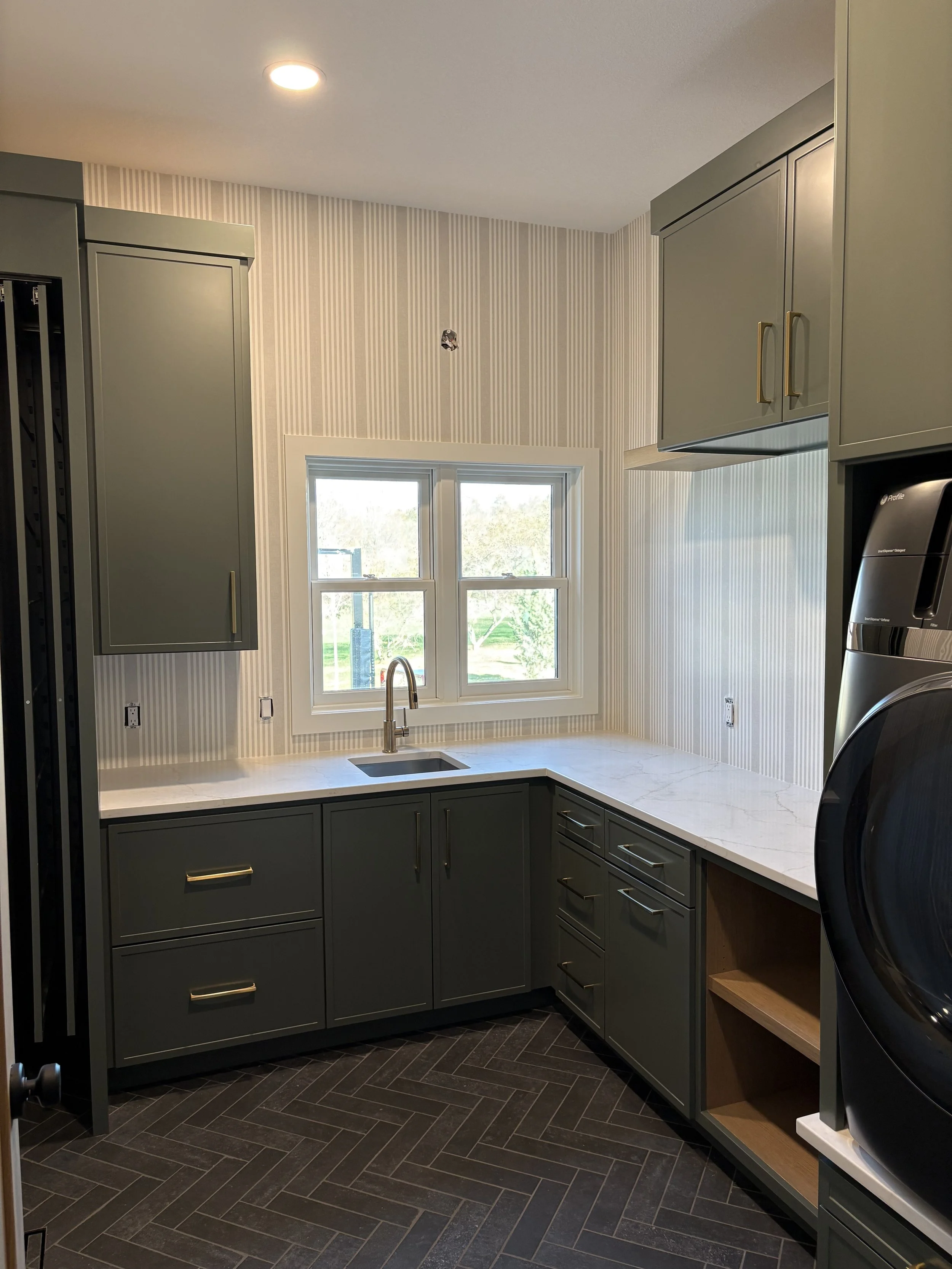 A modern kitchen with dark gray cabinets, white marbled countertops, a window above the sink, and black herringbone tile flooring.