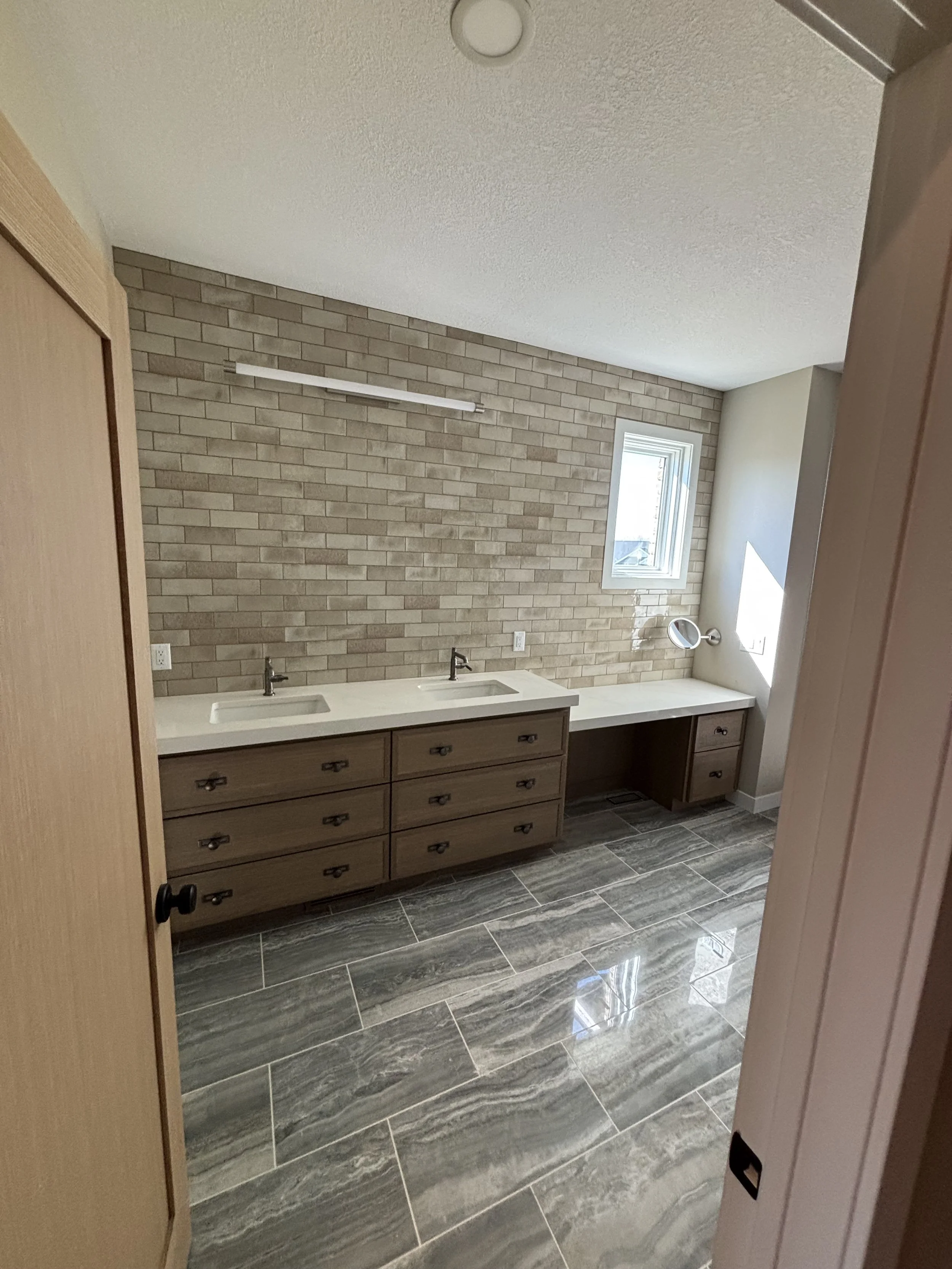 Bathroom with dual sinks, brick accent wall, small window, and gray tiled floor.