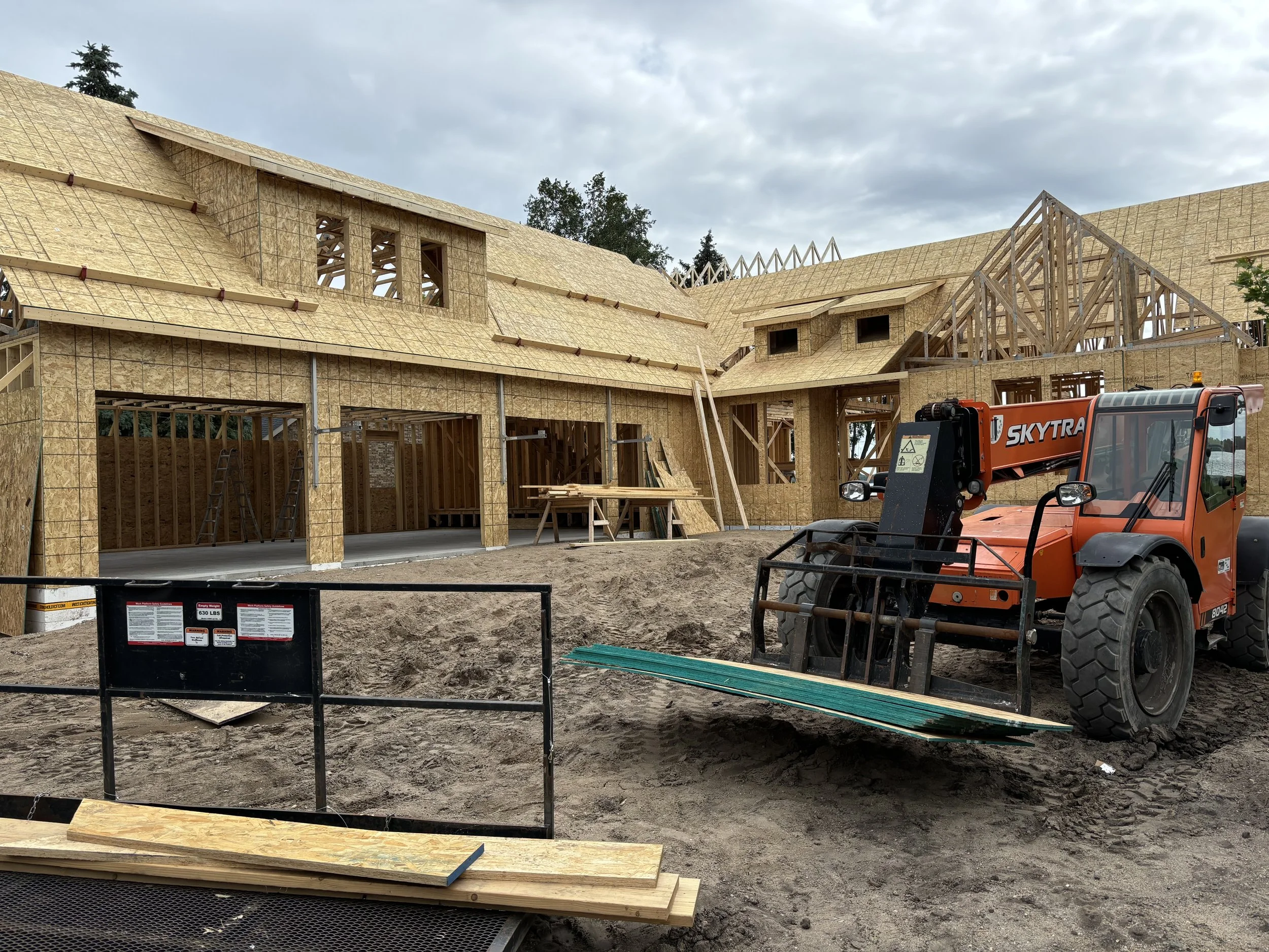 Residential building under construction with wooden framing and roofing. Construction equipment and materials are visible on site with cloudy sky overhead.