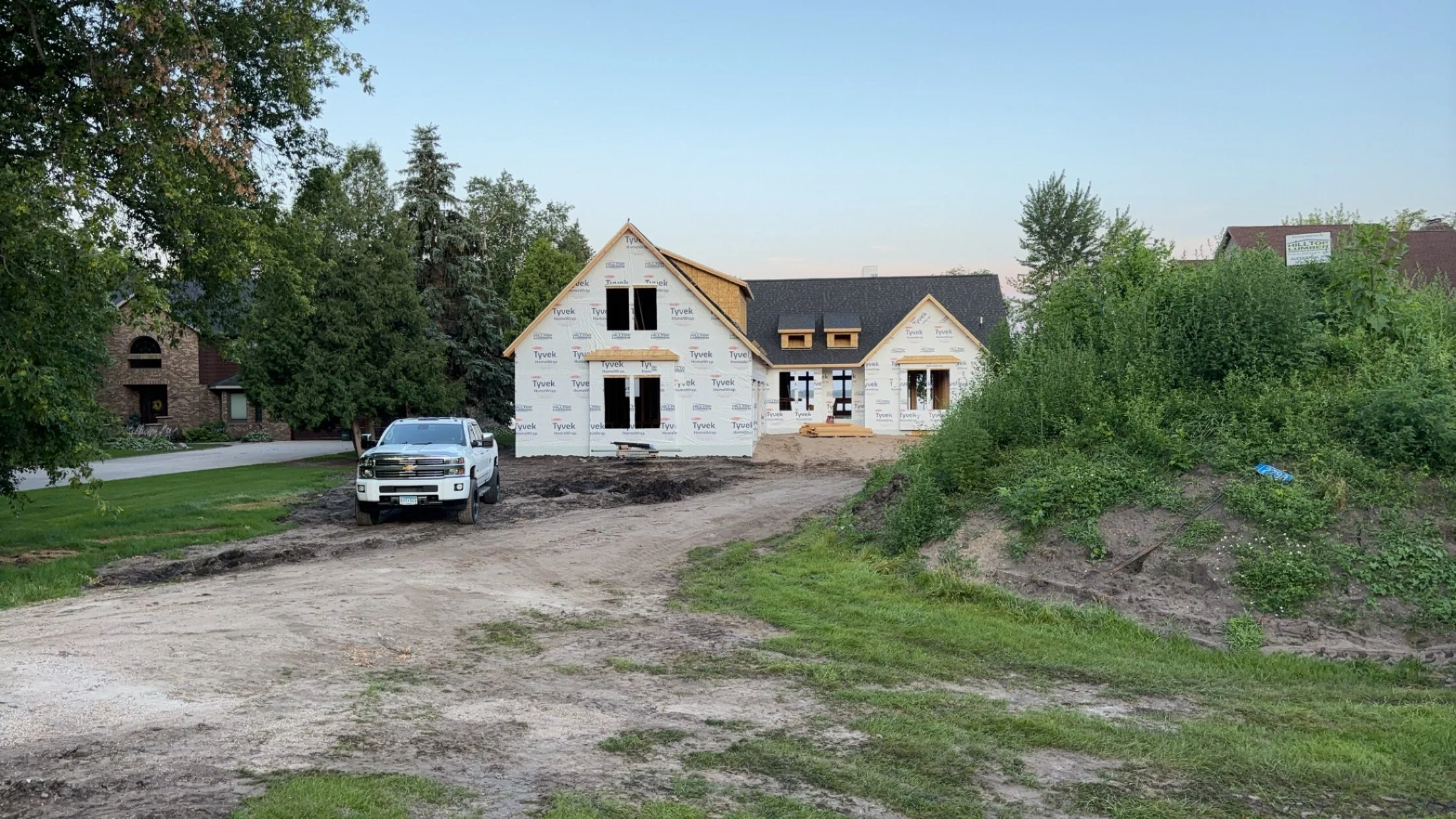 A house under construction with an unfinished exterior, surrounded by trees and a dirt driveway, and a white pickup truck parked in front.