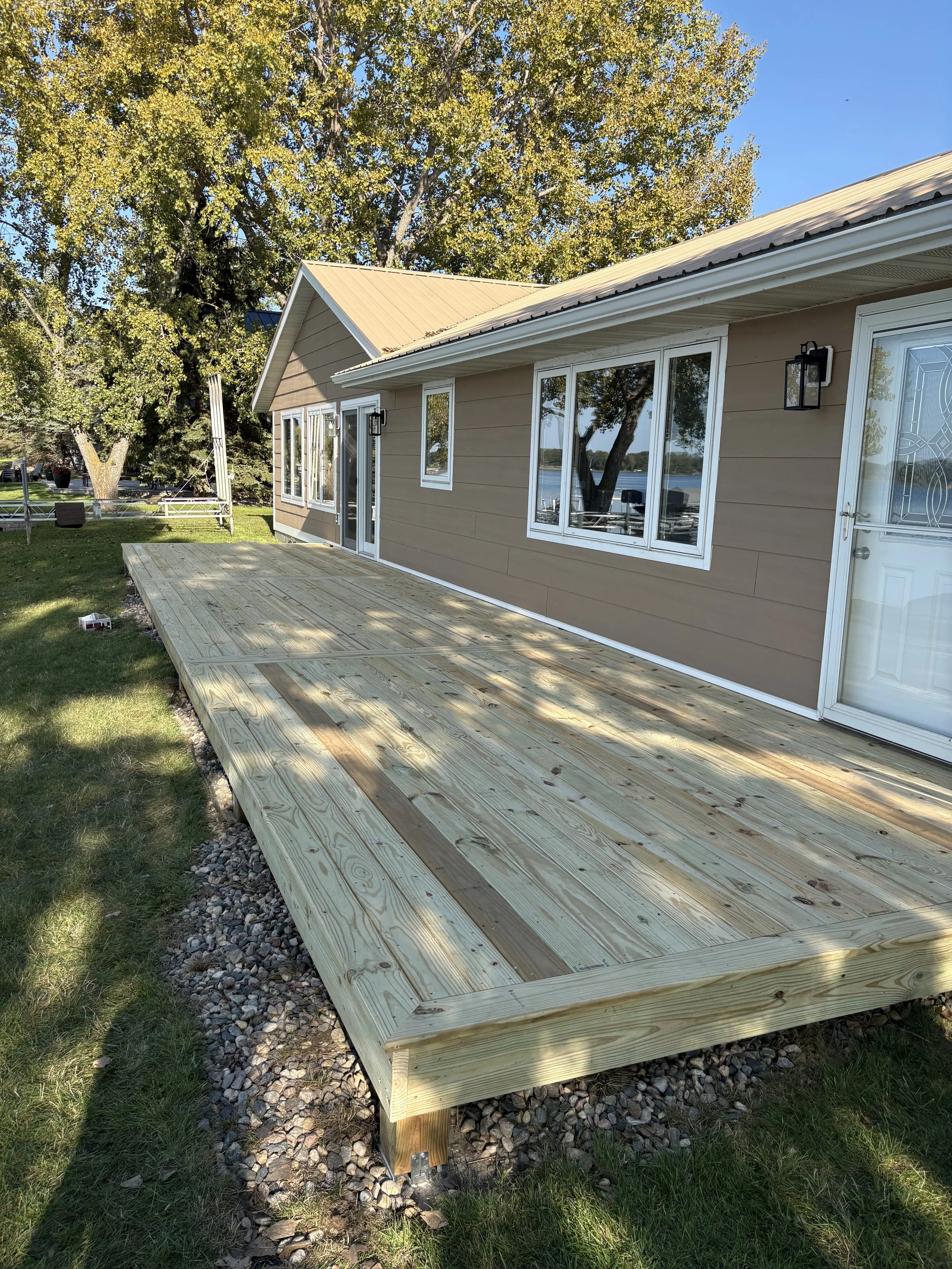 A newly built wooden deck attached to the house with a set of concrete steps leading to the yard, under a large tree with autumn foliage.