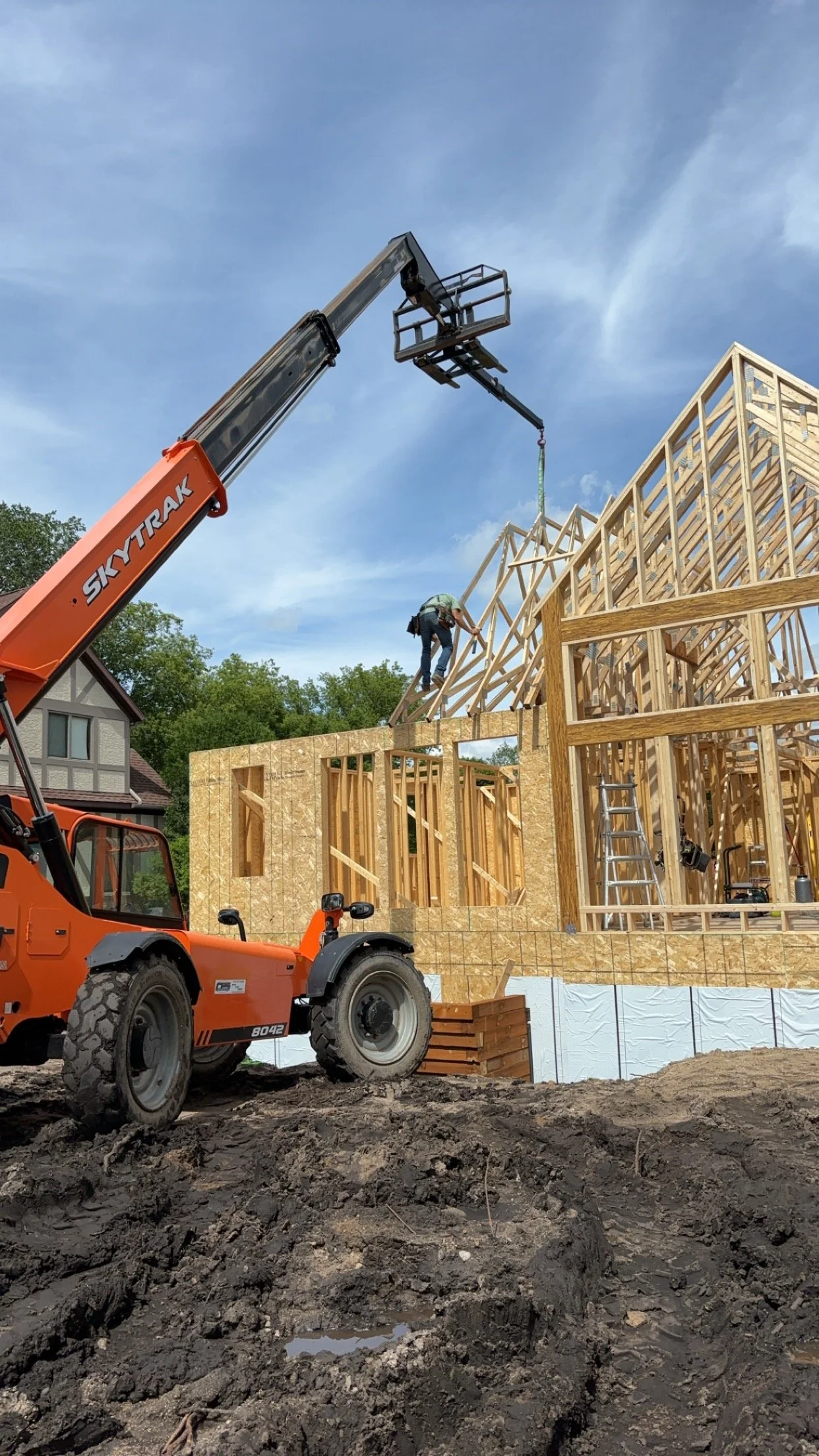 Construction site with a wooden house frame and a worker on the roof, using a crane to lift materials.